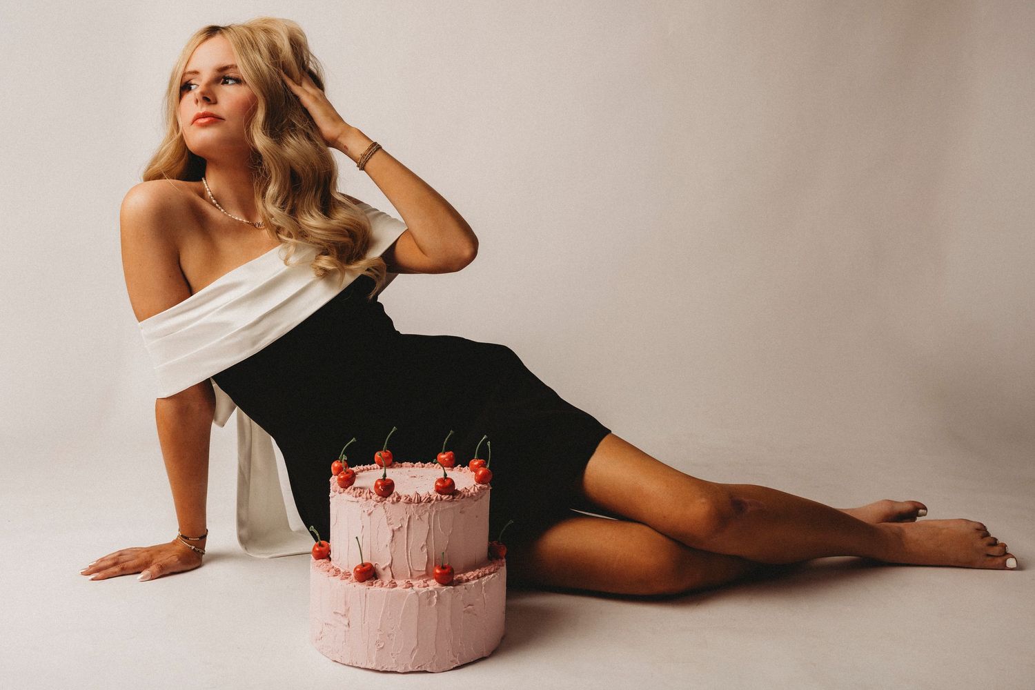 Blonde woman in black dress posing beside a two-tier pink birthday cake with cherries on white background. celebrating 18th birthday.