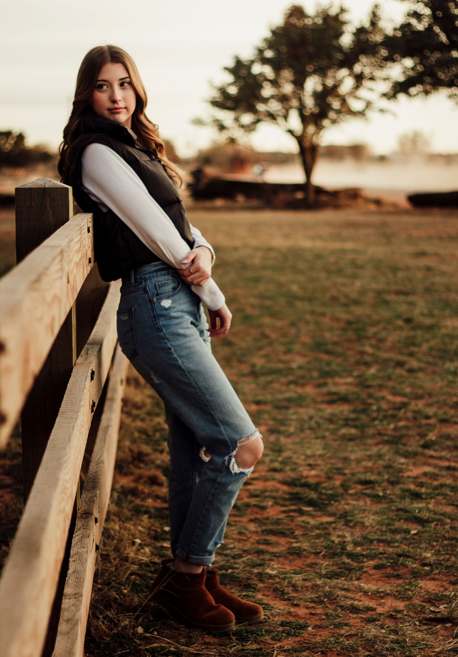 A person in denim overalls leans against a wooden fence during golden hour sunset in a rural field setting.