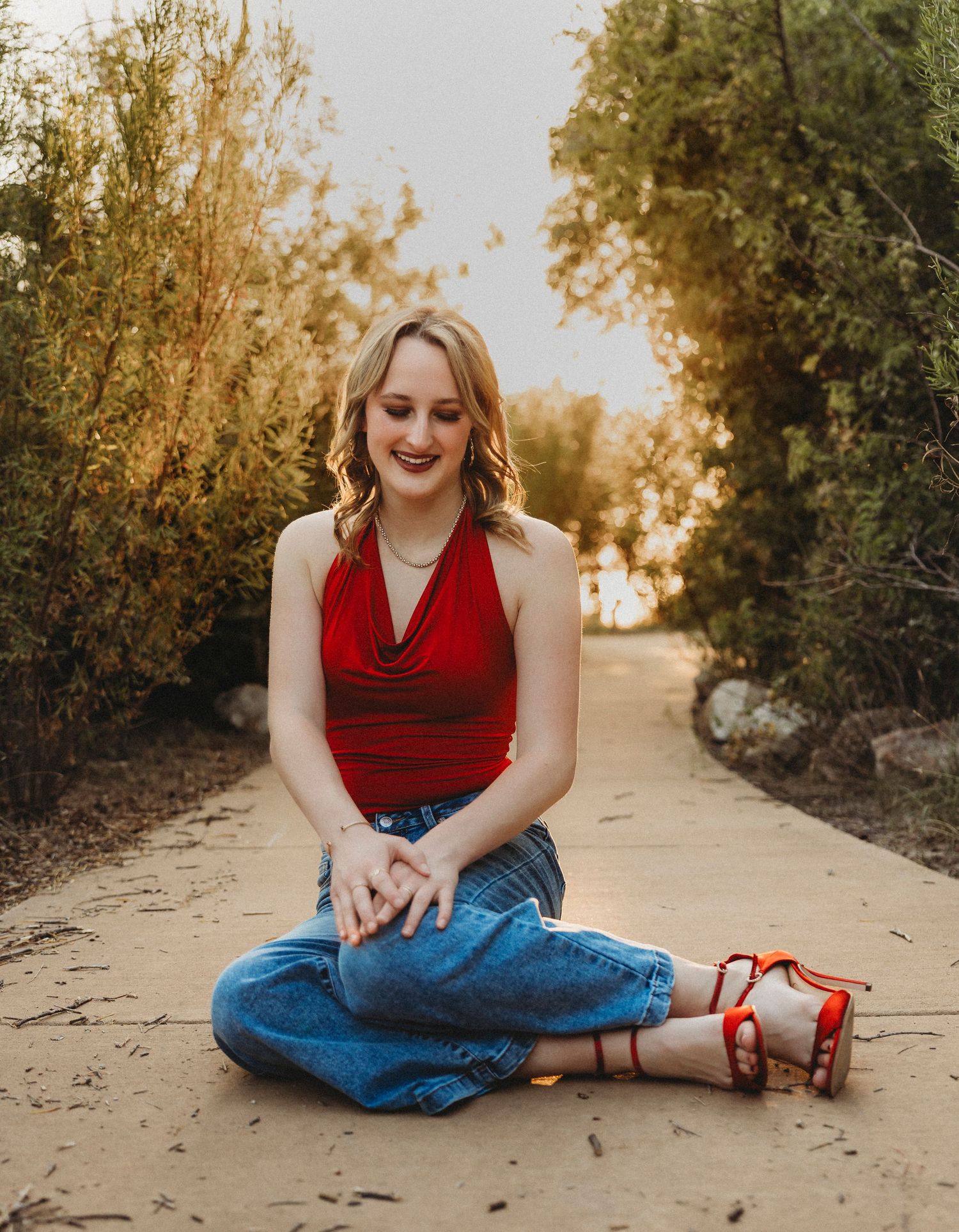 Smiling high school senior in a red top seated on a tree-lined pathway during a golden hour senior portrait session in Abilene, Texas.