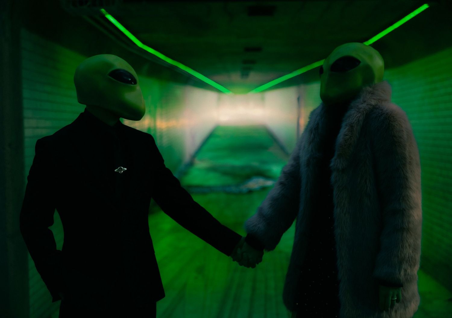 Two figures in alien masks shake hands under green laser lights in a dramatic, sci-fi themed scene.