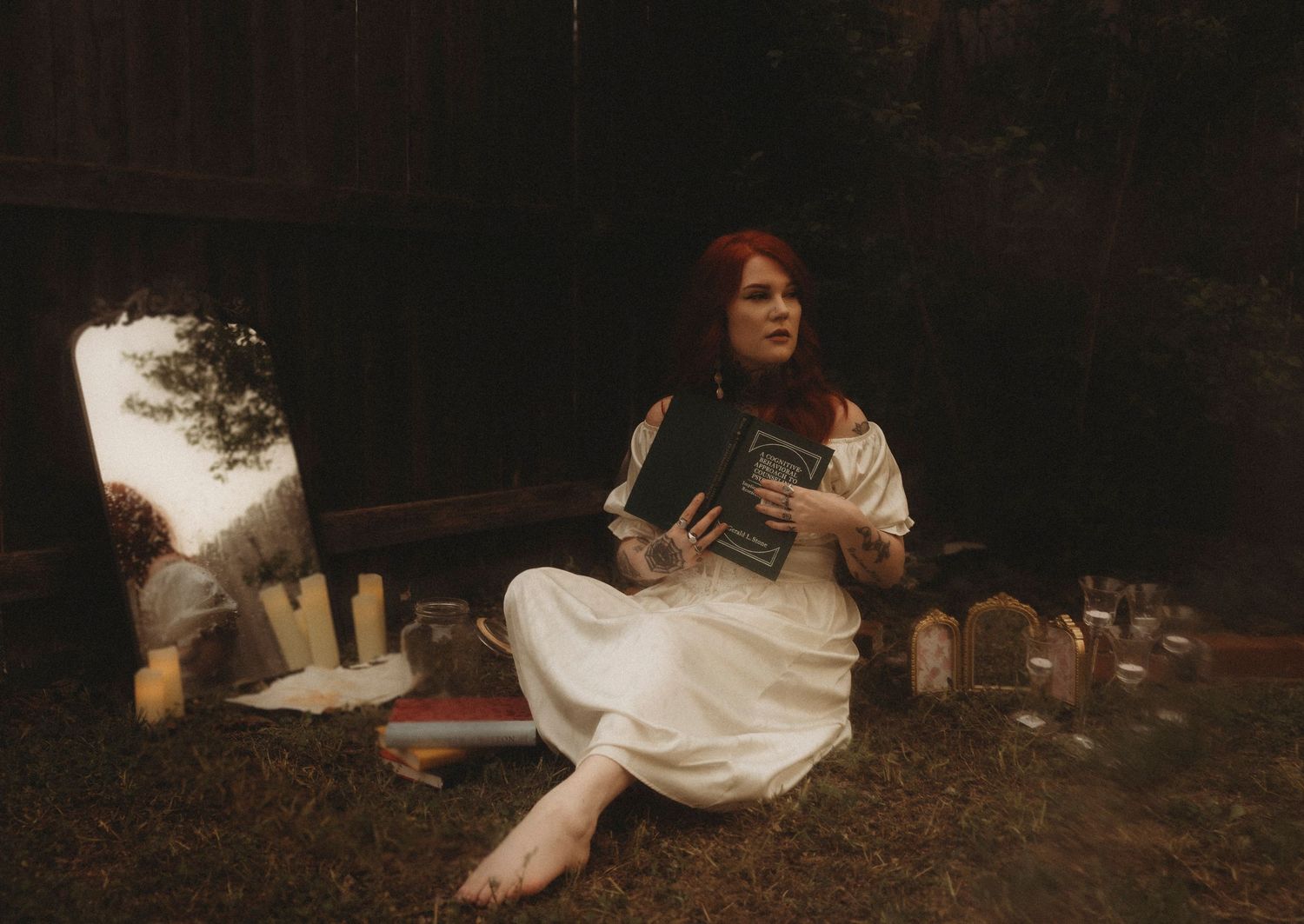 Red-haired woman in white dress sitting outdoors holding a book, surrounded by candles and mystical items.