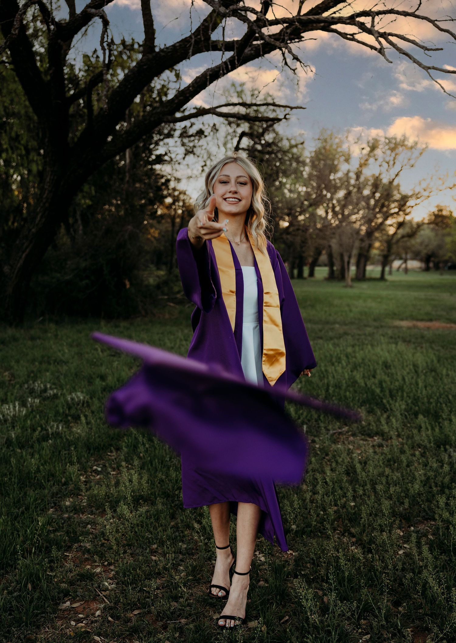 Wylie High School Graduate in purple cap and gown twirls joyfully in a grassy field at sunset during a graduation photoshoot.