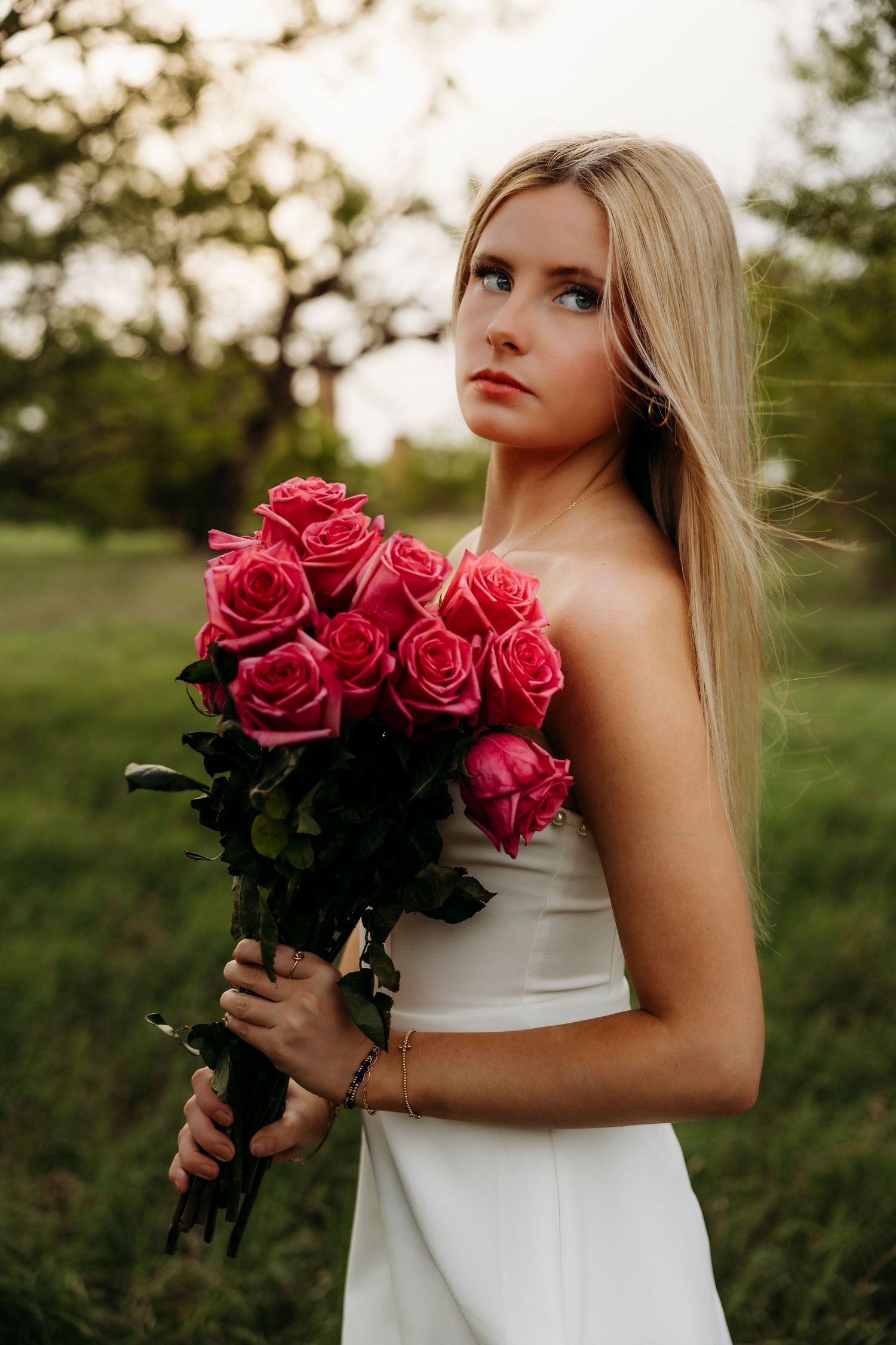 High School Senior in white dress holding bouquet of pink roses outdoors at dusk.