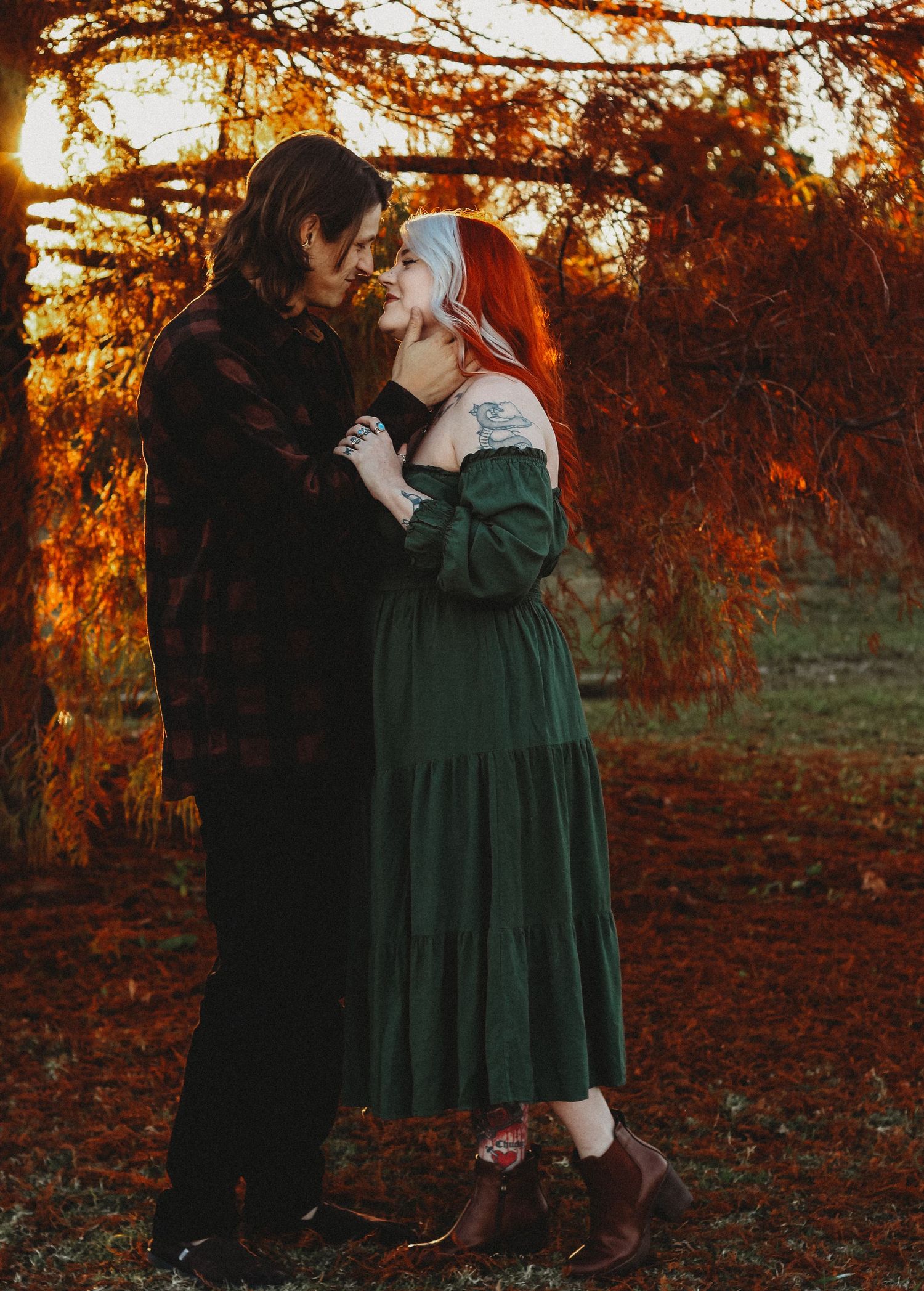 Couple in romantic autumn embrace, woman in green dress with red hair, man in dark coat, golden sunset foliage backdrop.