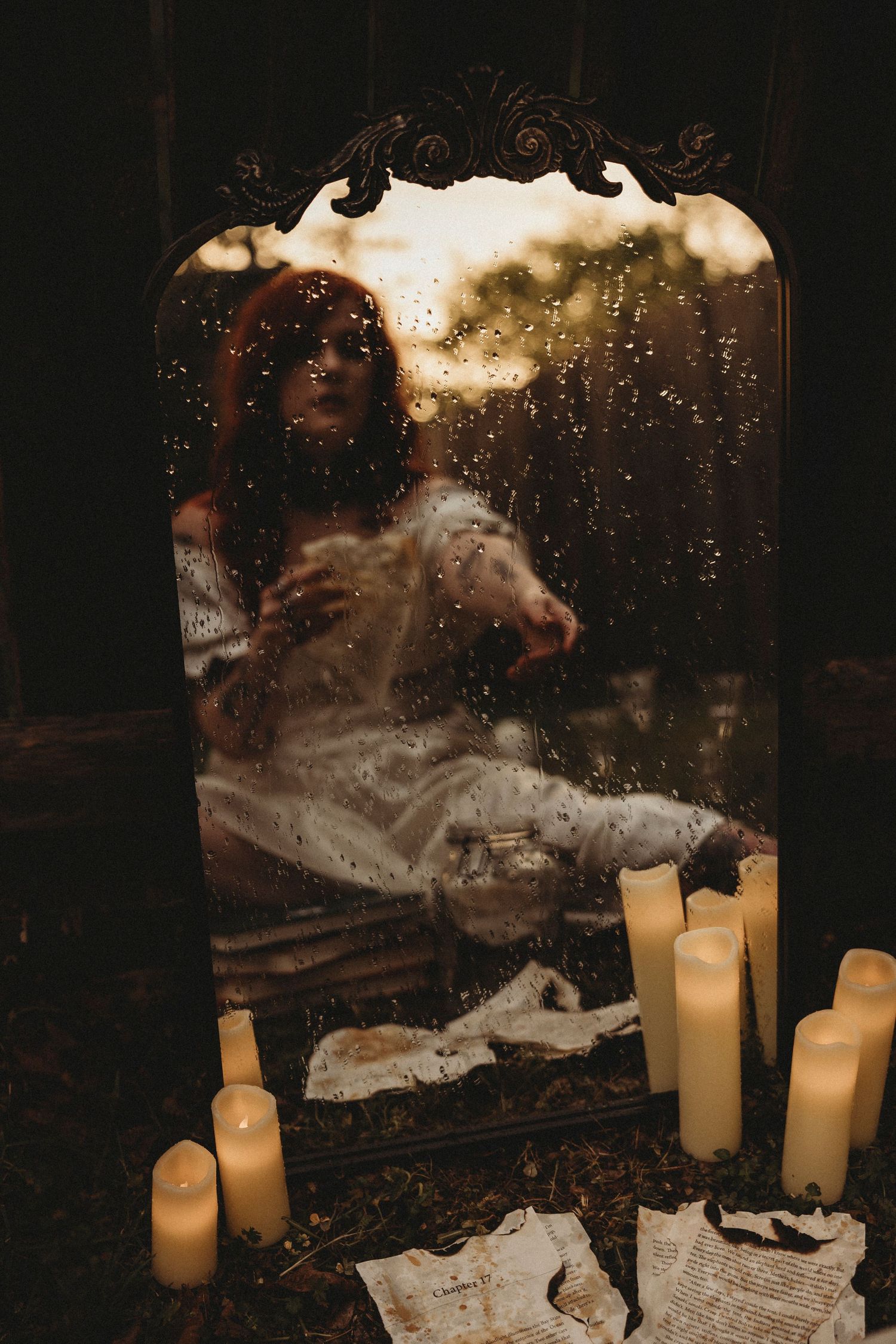 Woman in white dress reflected in ornate mirror surrounded by glowing candles and scattered papers in dark setting.