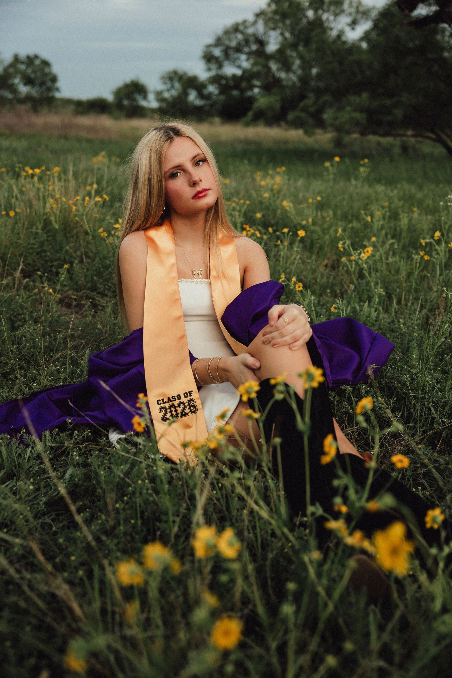 A high school senior poses in a wildflower field wearing a purple gown and gold Class of 2026 graduation stole.