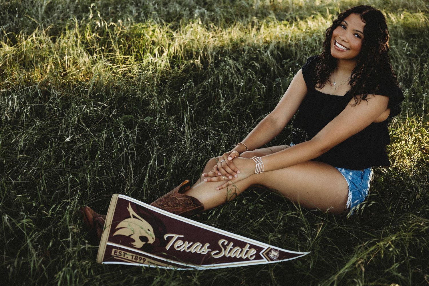 High school senior sitting in tall grass with a Texas State University pennant during a college-bound senior session in Abilene, Texas