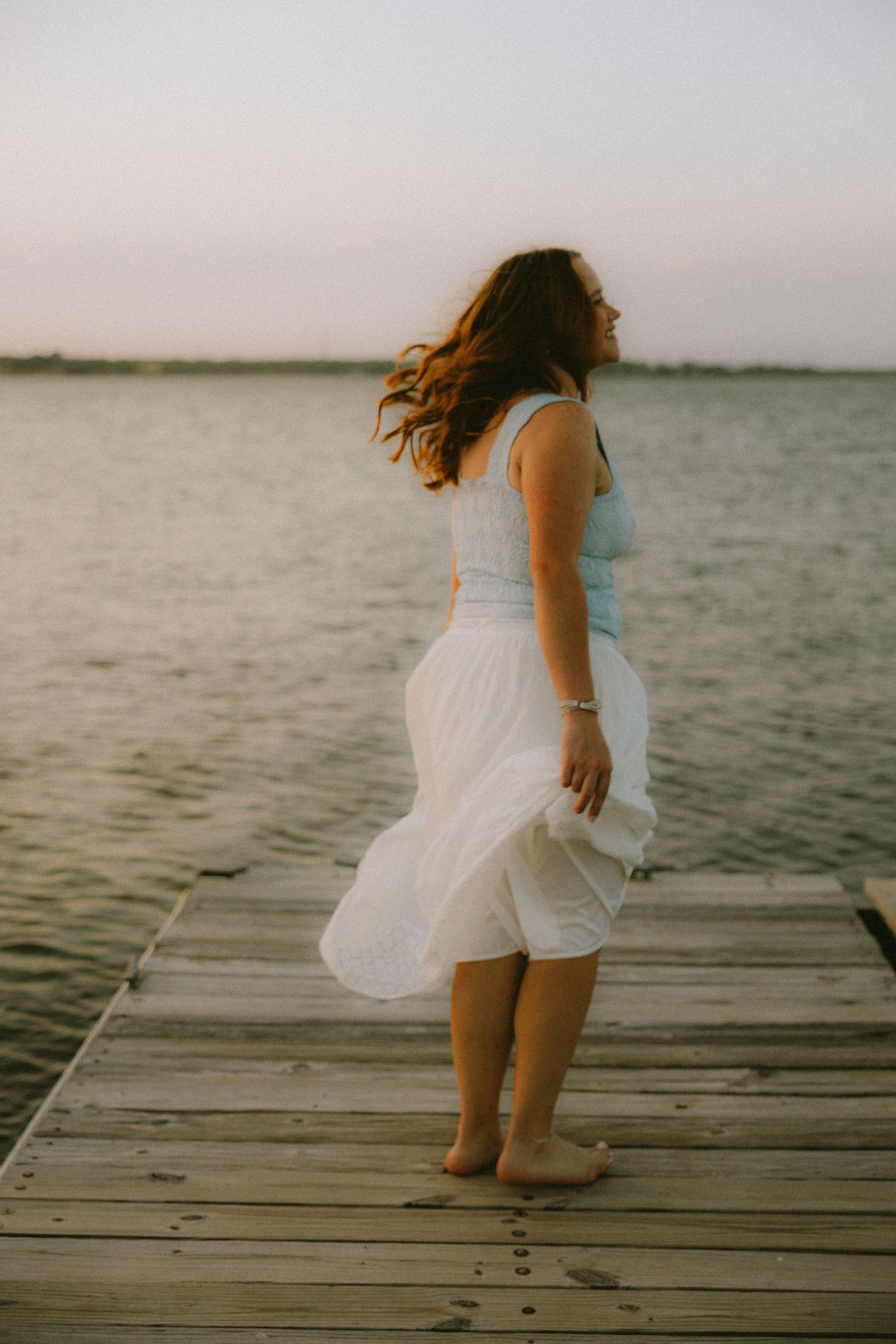 Abilene High School Senior in white skirt standing barefoot on wooden dock by calm water at sunset with hair blowing in wind.