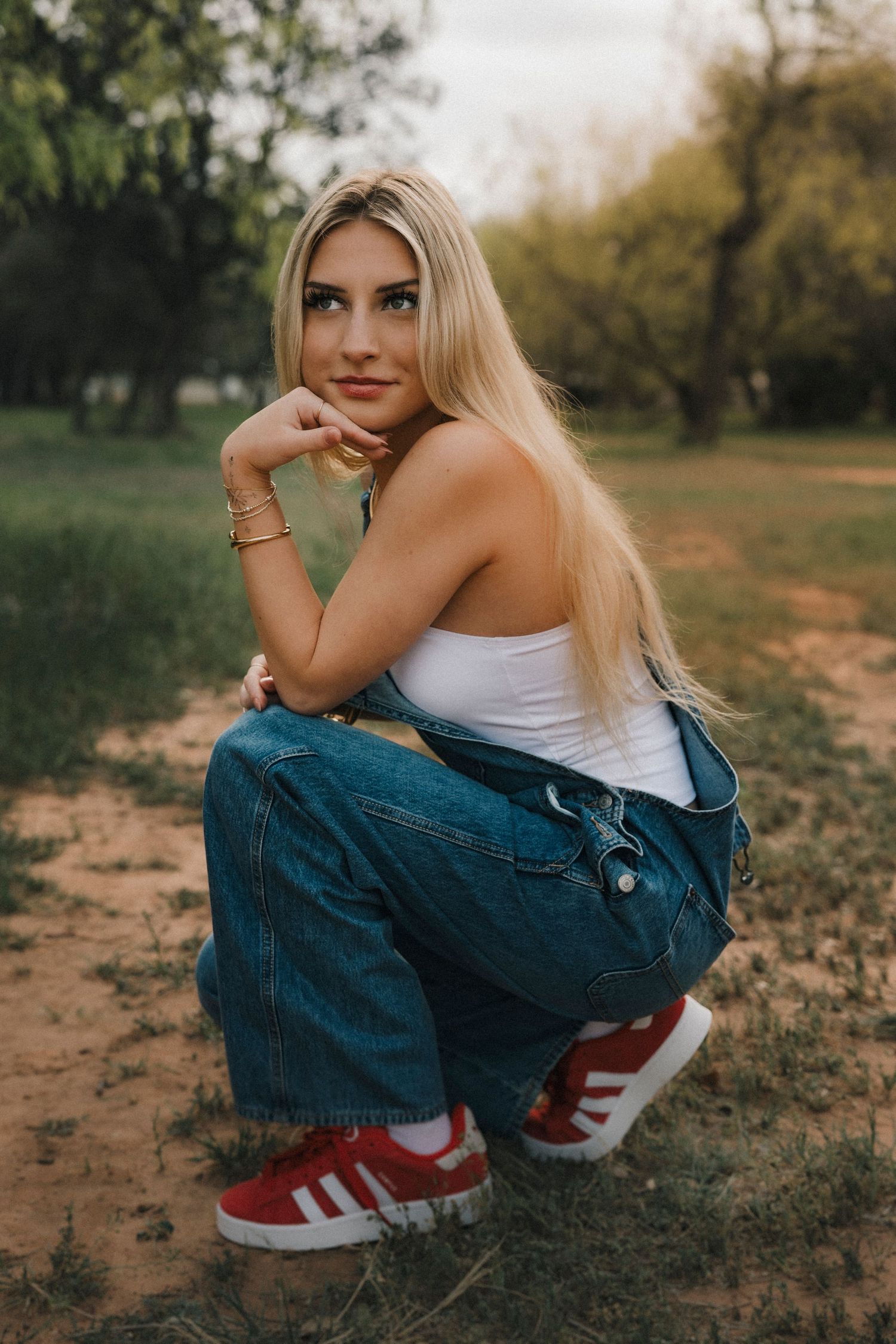 Senior girl  in denim overalls and red Adidas sneakers crouching outdoors in a park setting. Abilene Texas