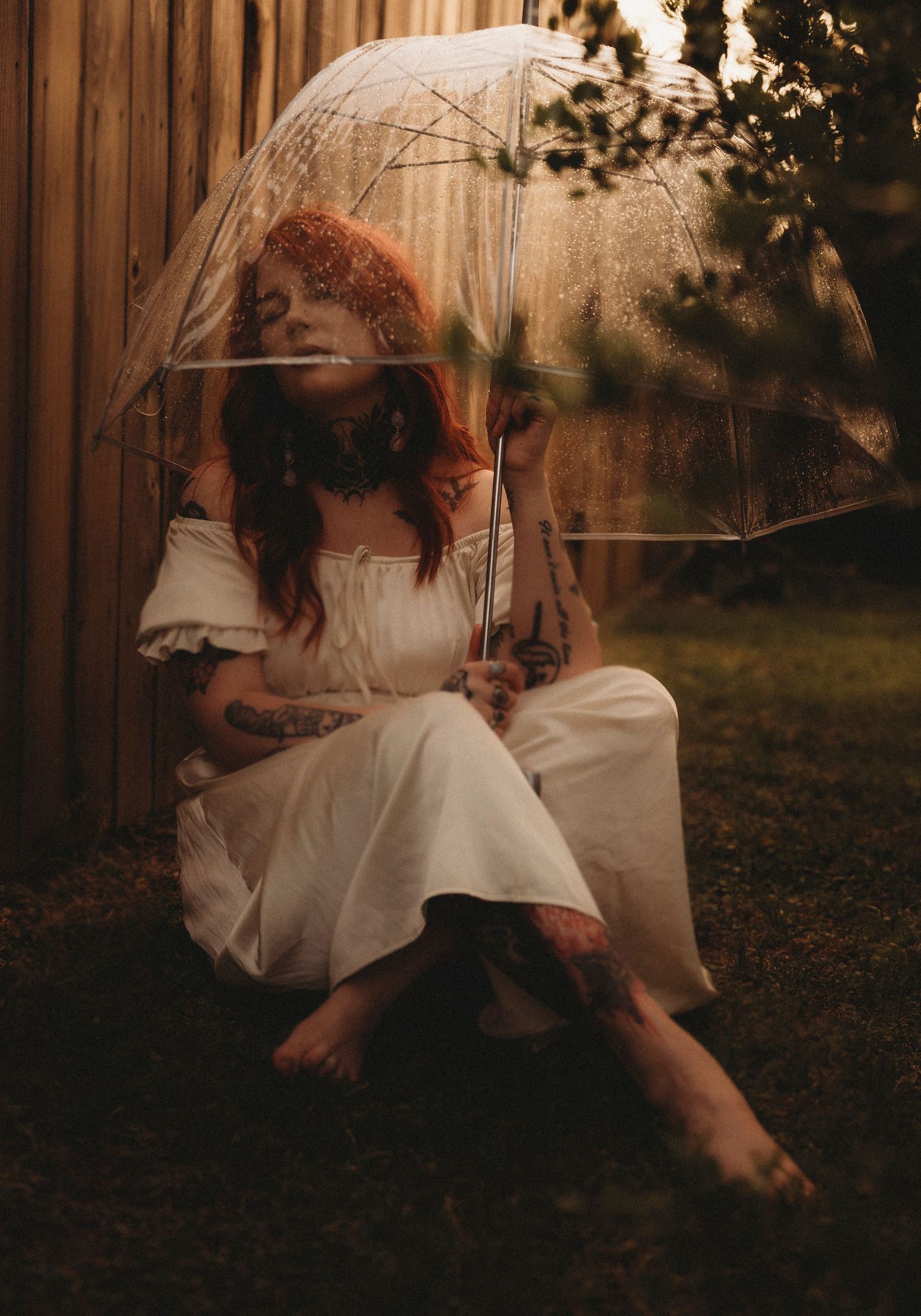 Tattooed redhead woman in white dress sitting barefoot on grass holding clear bubble umbrella outdoors at dusk.