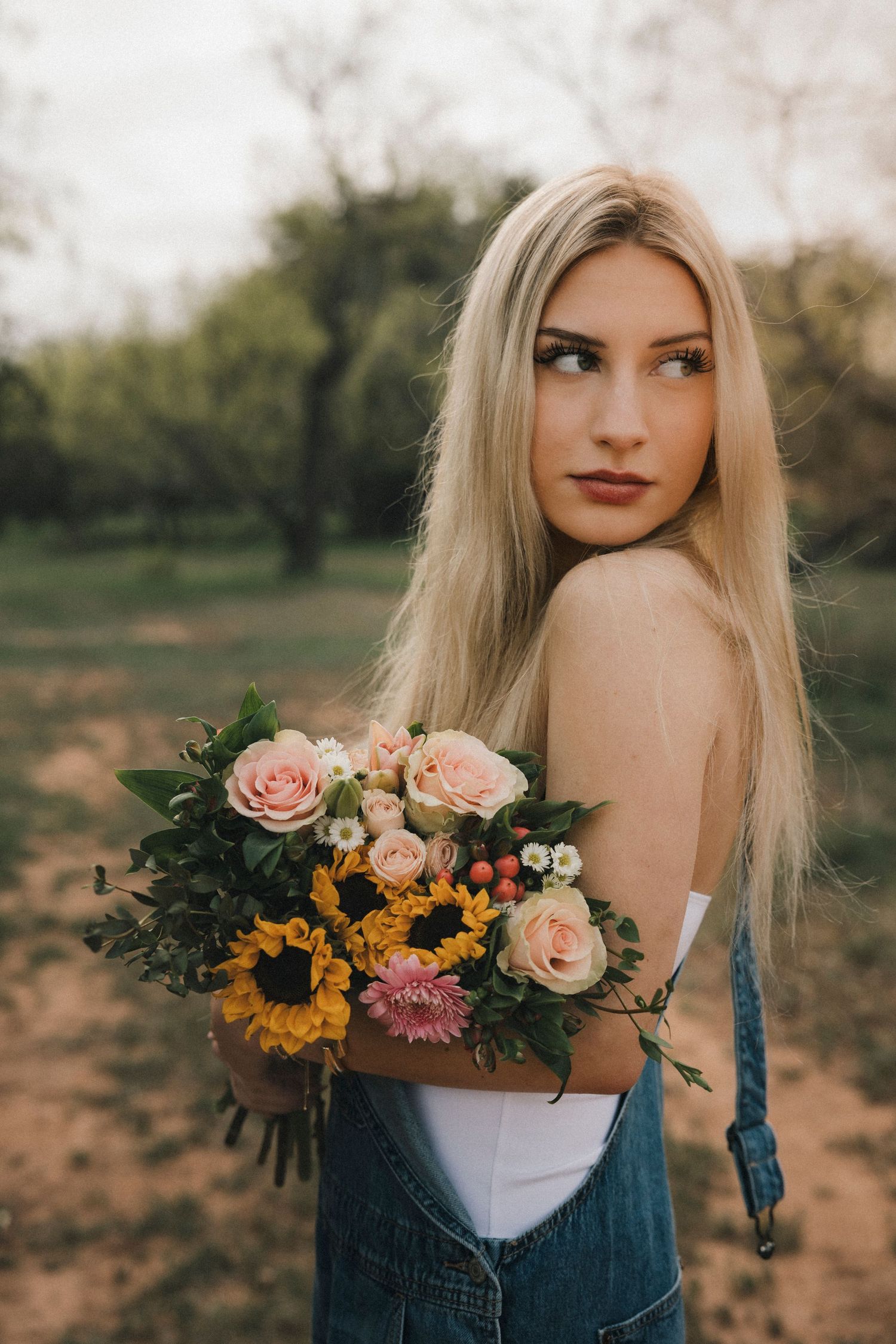Senior High School Girl holding colorful bouquet of sunflowers and roses, wearing denim overalls outdoors.