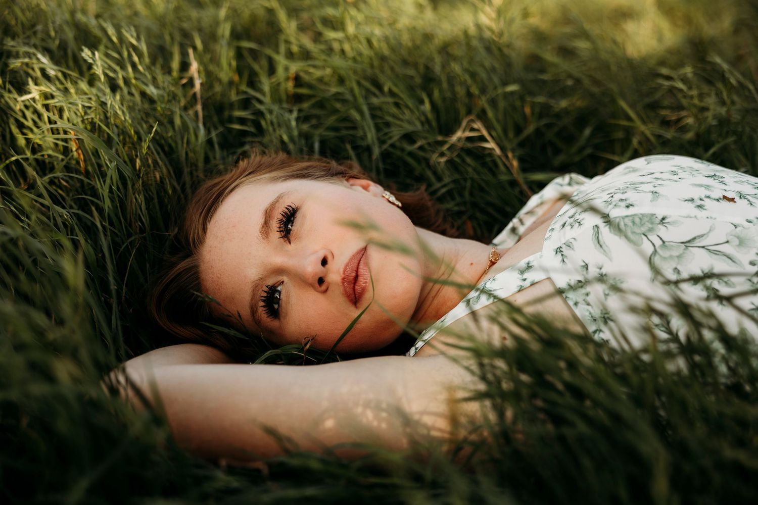Abilene TX High School senior in a floral dress lying peacefully in tall green grass, gazing upward in soft natural light.