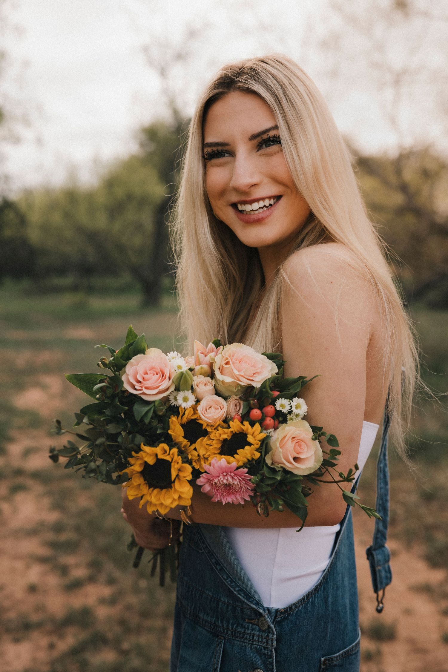 Senior girl photoshoot holding a colorful bouquet of sunflowers and roses outdoors in a white top and denim overalls.