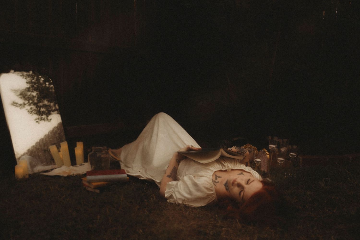 Woman in white dress lying on grass at night surrounded by candles and glowing light in a dreamy outdoor scene.