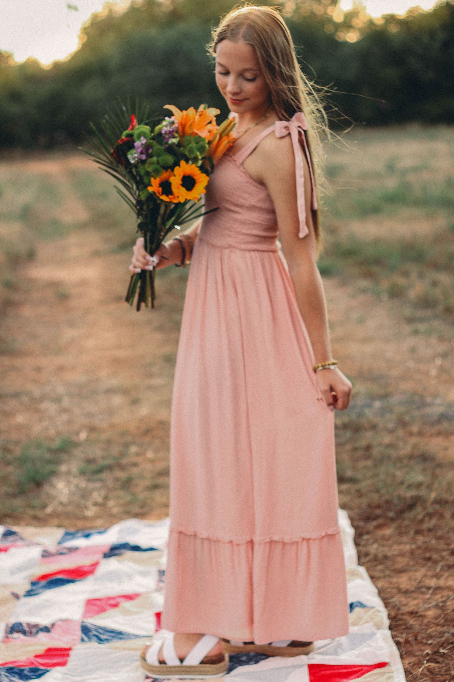 High school senior holding a colorful bouquet while wearing a pink dress in a field at sunset.