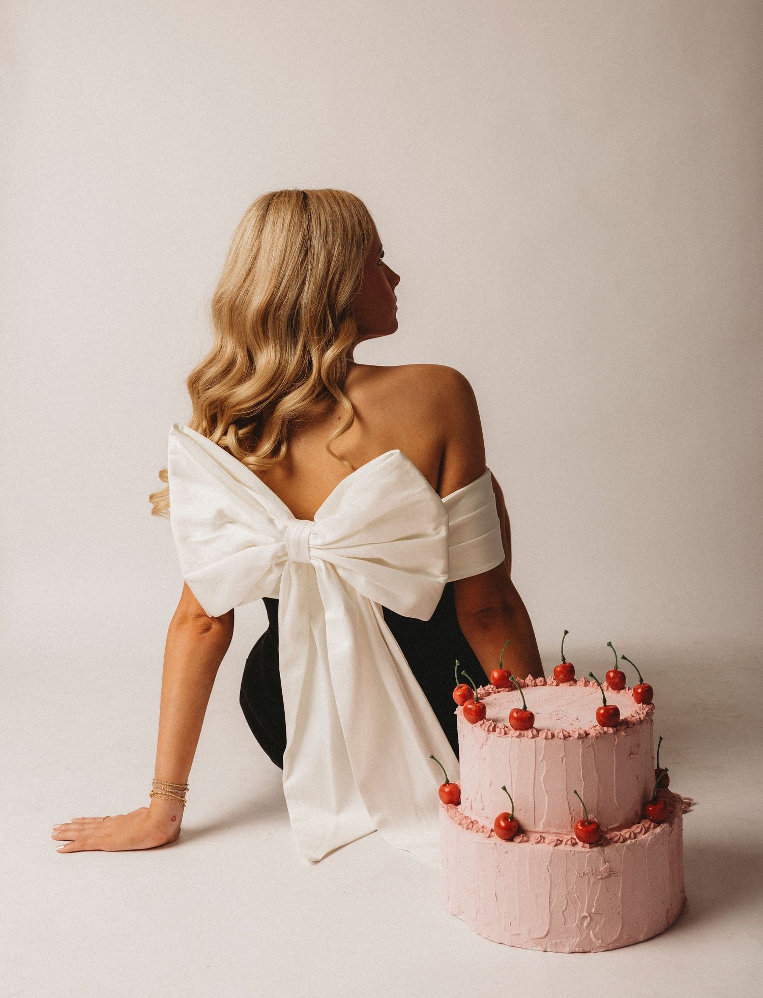 Blonde woman in black dress with white bow sits beside a pink two-tier birthday cake topped with cherries.