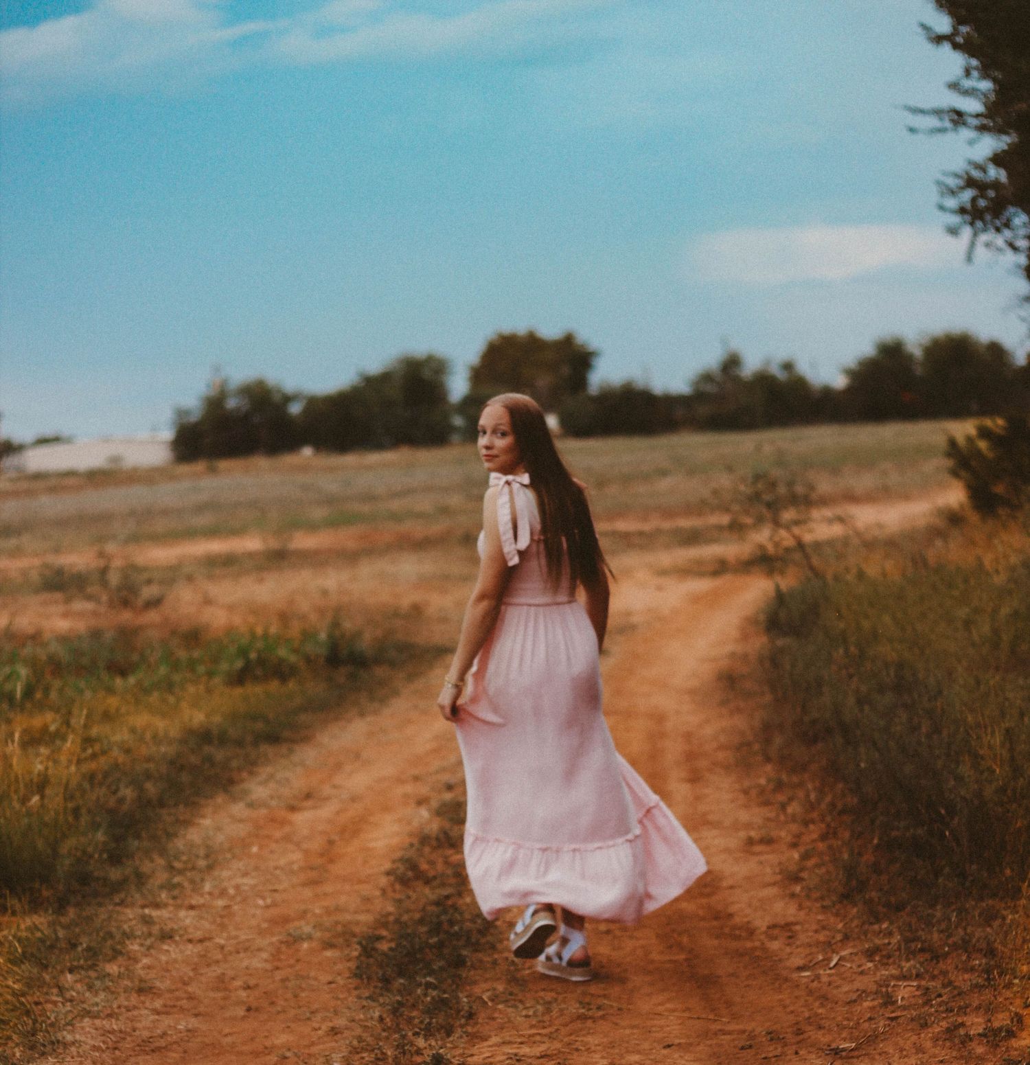 Person in flowing pink dress walks along a winding dirt path through golden fields at sunset.