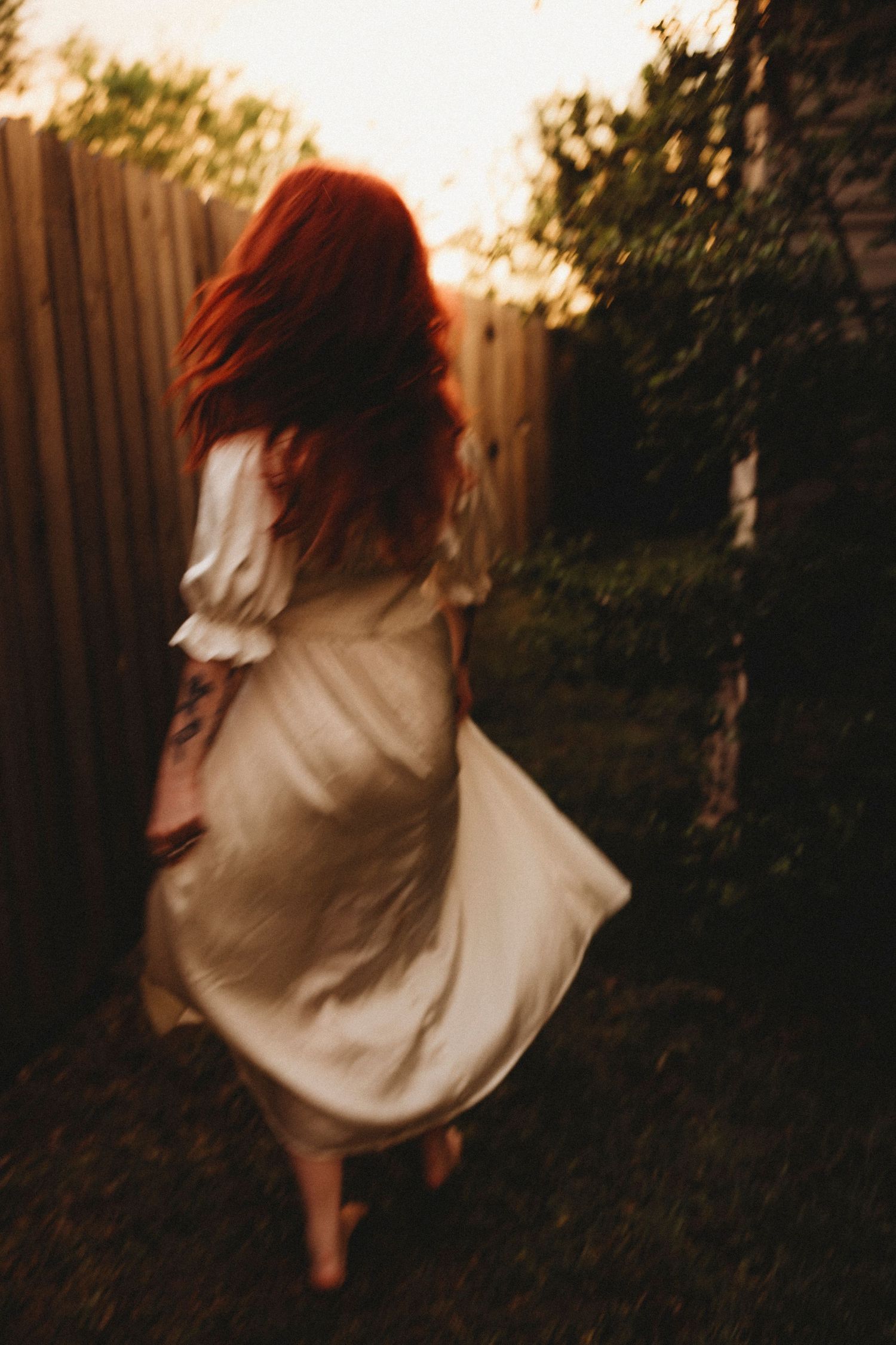 Red-haired woman in white dress walking through garden at sunset.