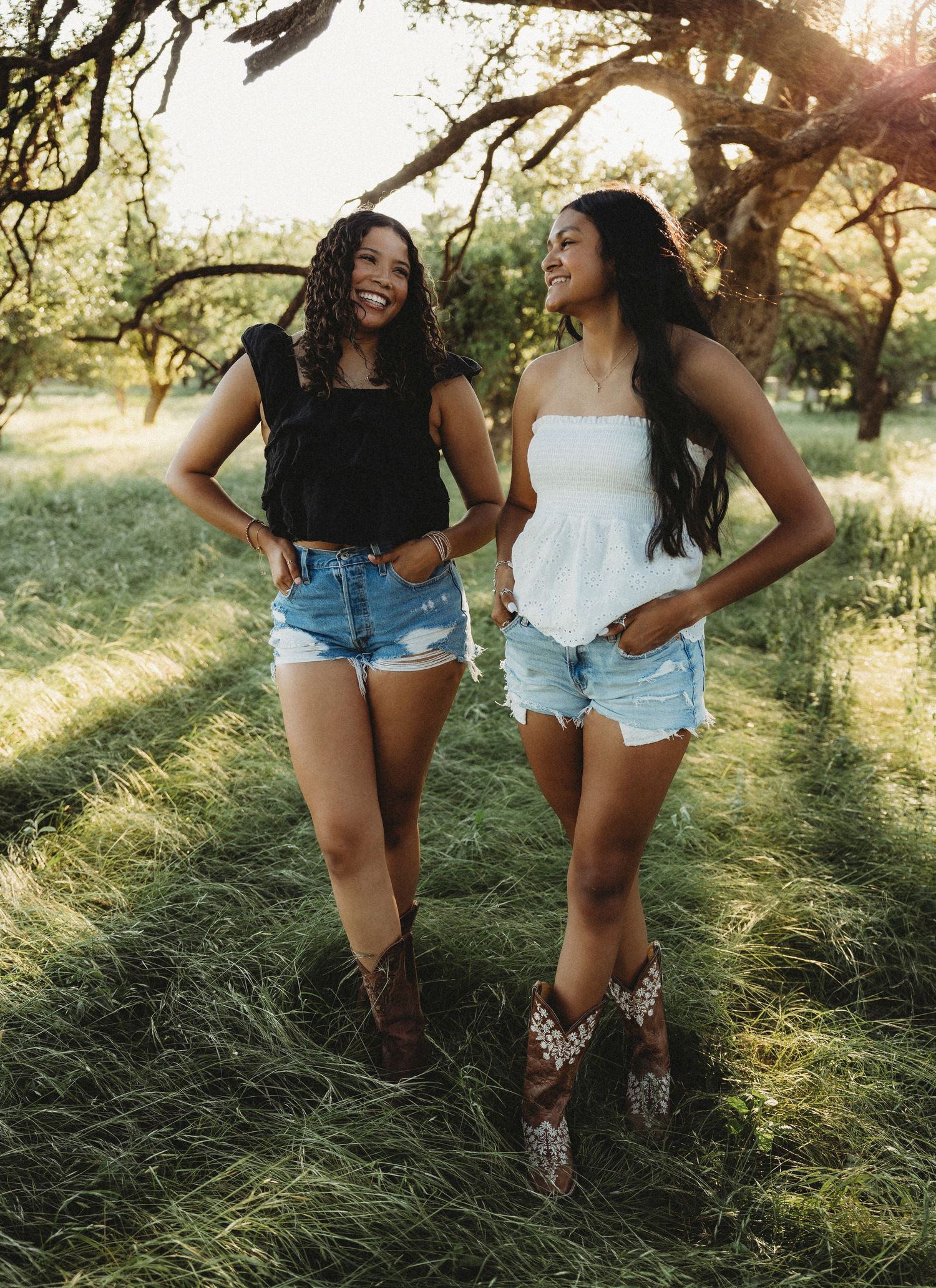 Two smiling best friends in denim shorts and boots pose outdoors in a sunlit grassy field with trees.