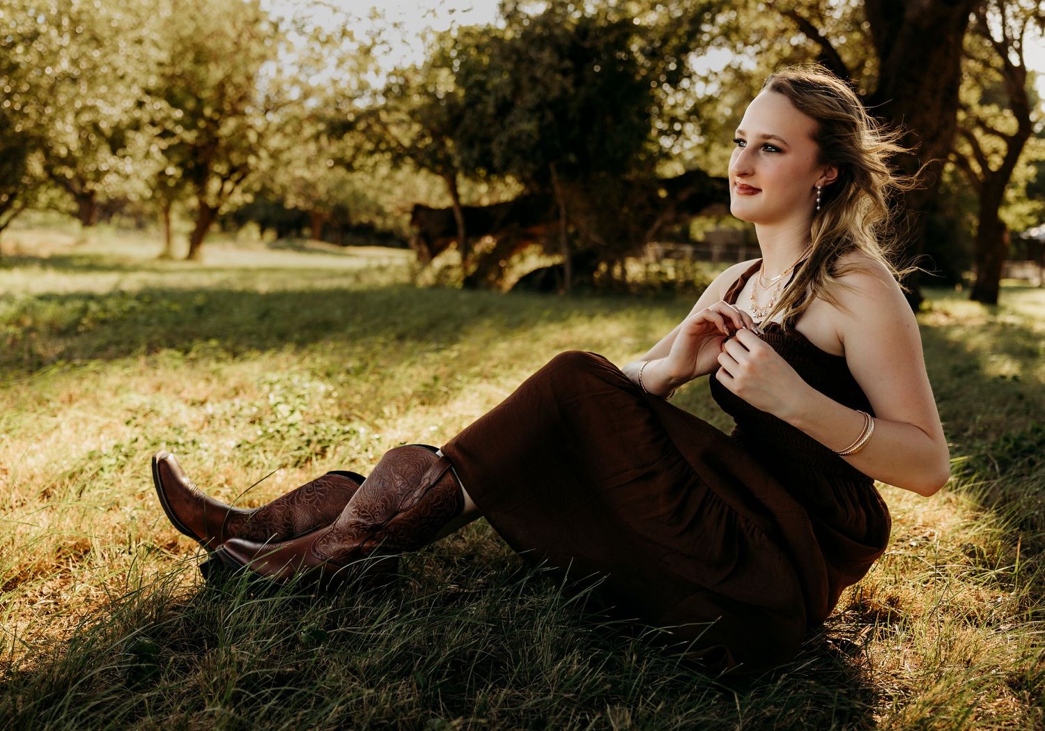 Golden hour senior portrait of a high school senior sitting beneath oak trees in a rustic brown outfit during an outdoor session in Abilene,
