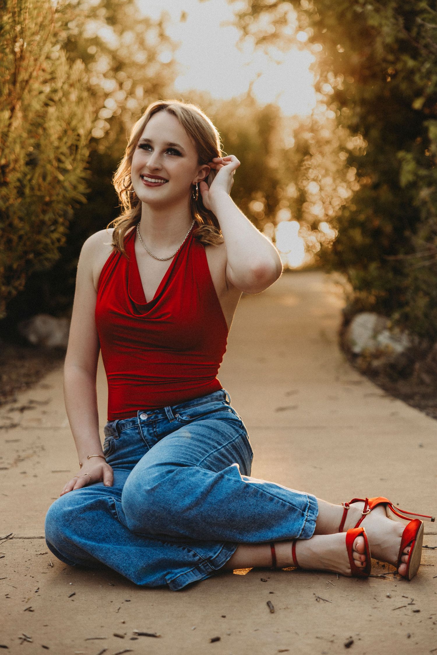 High school senior sitting on a tree-lined pathway in a red top and jeans during a golden hour senior portrait session in Abilene, Texas.