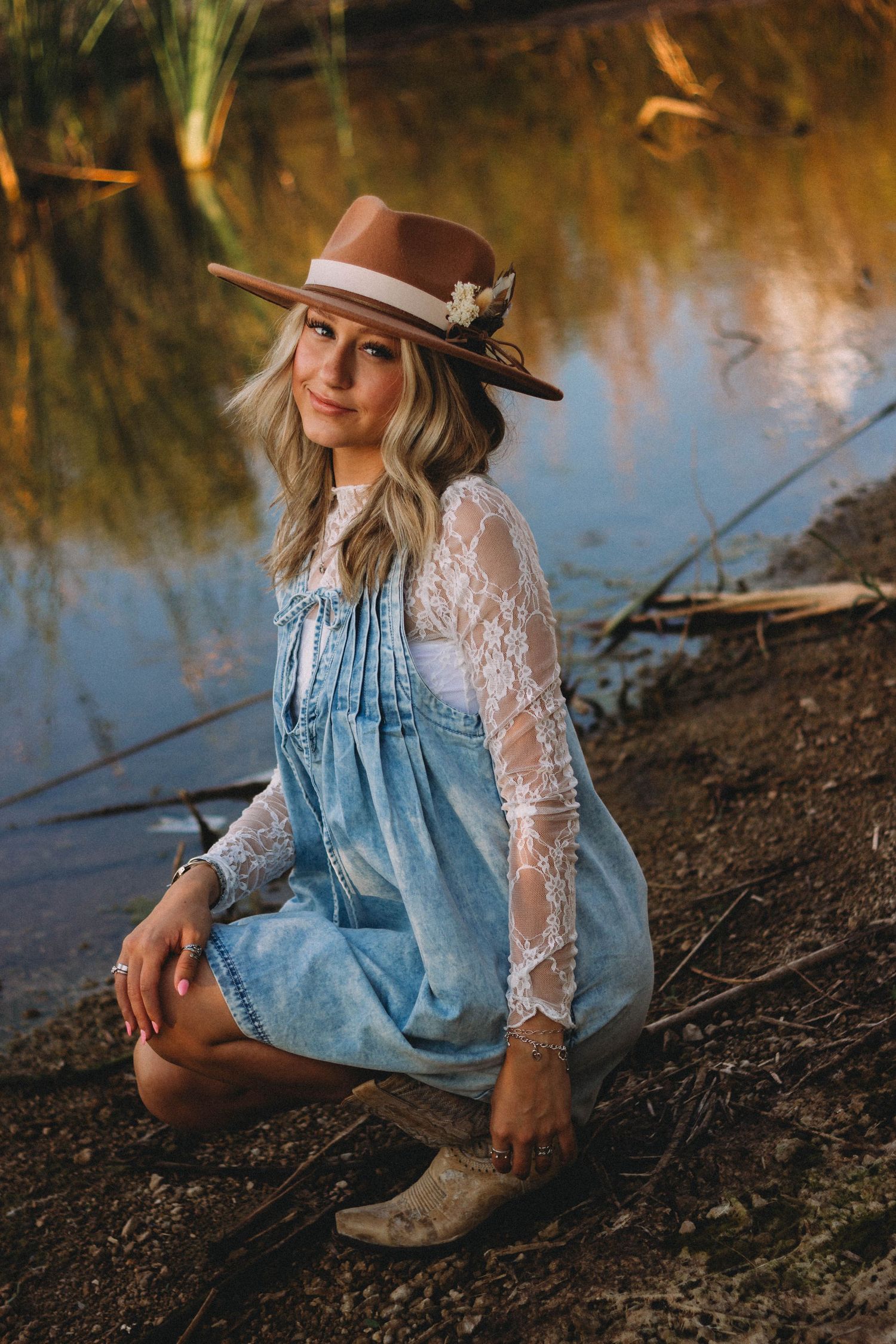 A dreamy sunset portrait of someone wearing a denim dress, lace sleeves, cowboy boots and hat sitting by a lake.