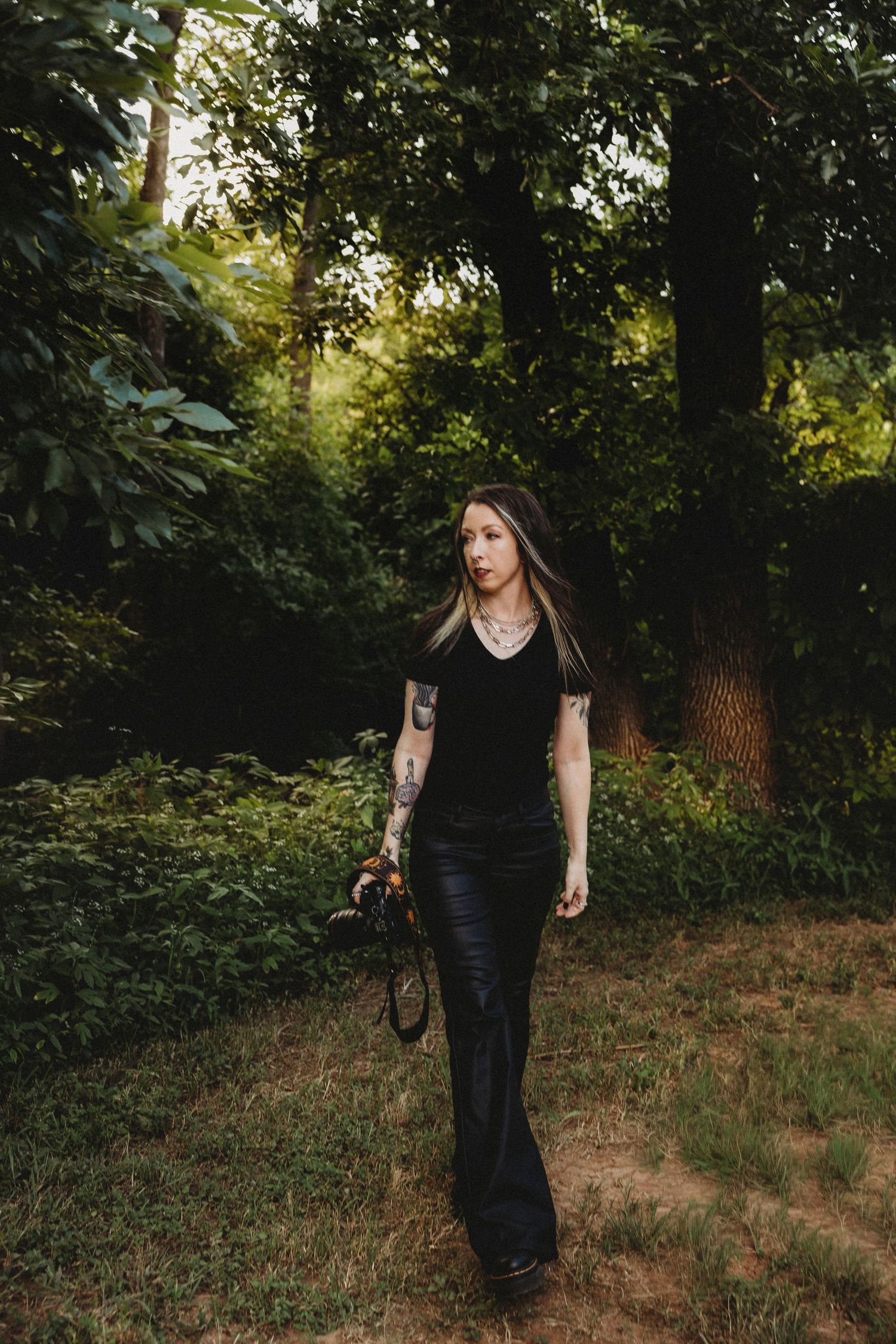 Tattooed photographer woman in black outfit walking through lush green forest, holding camera, golden hour light filtering through trees.