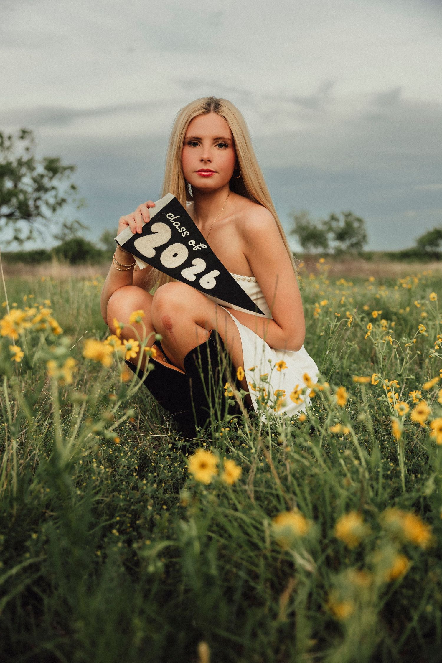 Blonde high school senior girl sitting in a wildflower field holding a black Class of 2026 pennant flag outdoors.