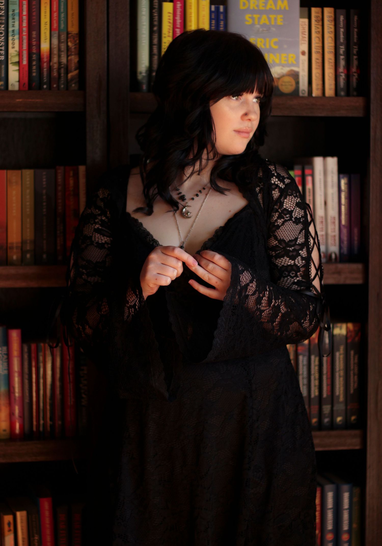 Senior girl in a black lace dress standing in front of a bookshelf during a moody editorial portrait session.