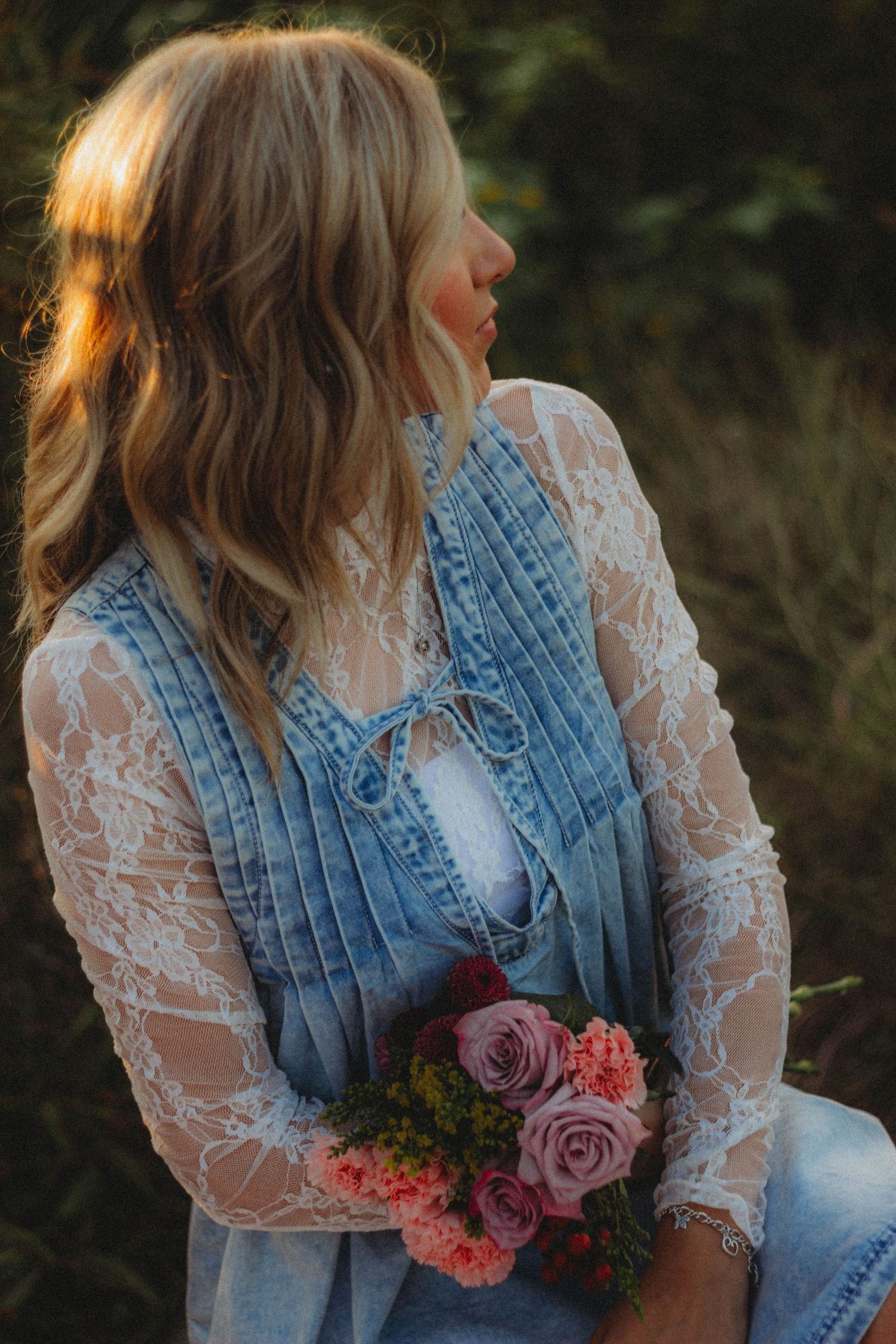 A person in a denim vest and lace sleeves holds a bouquet of pink roses in a romantic outdoor setting at sunset.