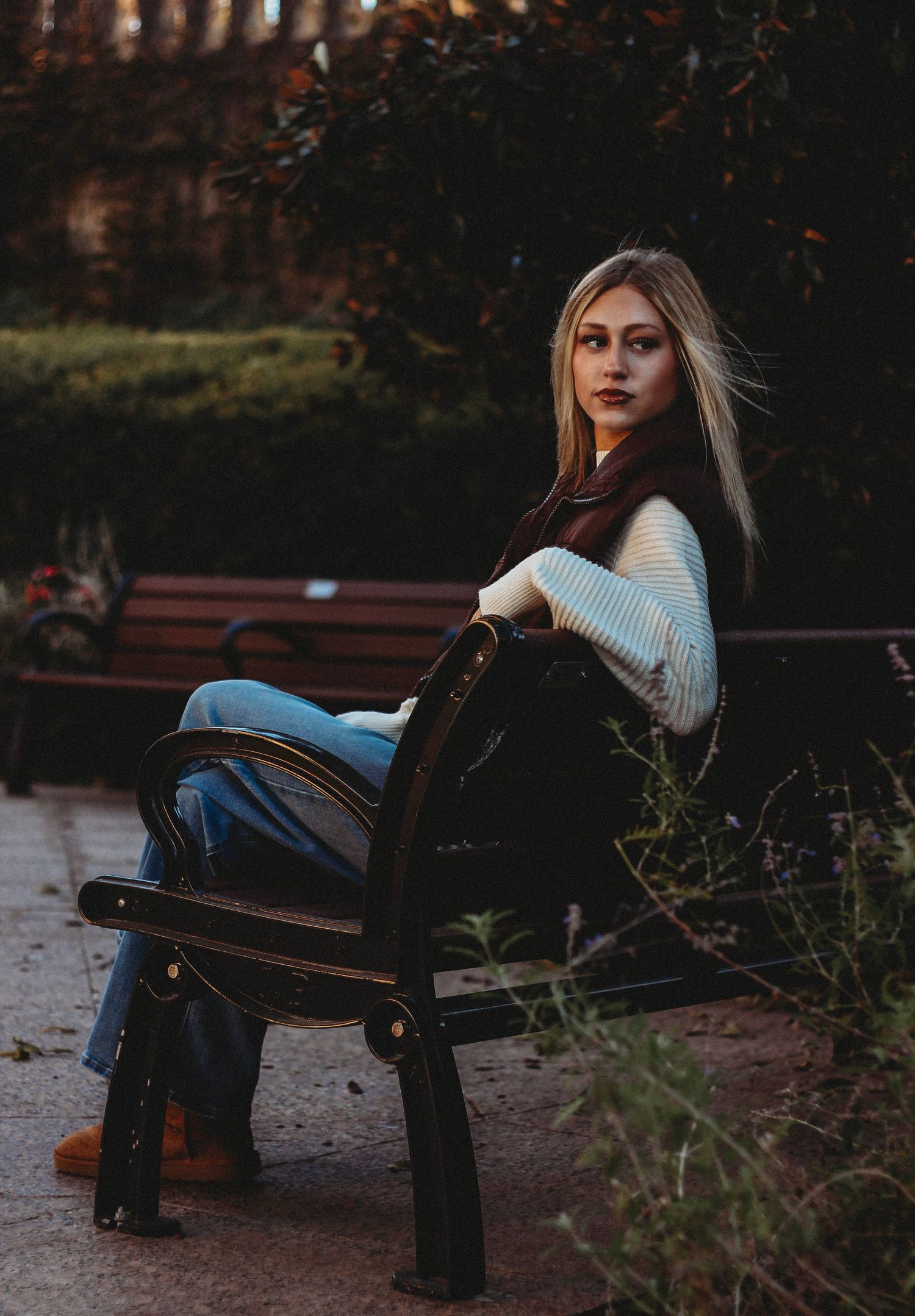A person in a white sweater and jeans sits thoughtfully on a park bench at dusk.