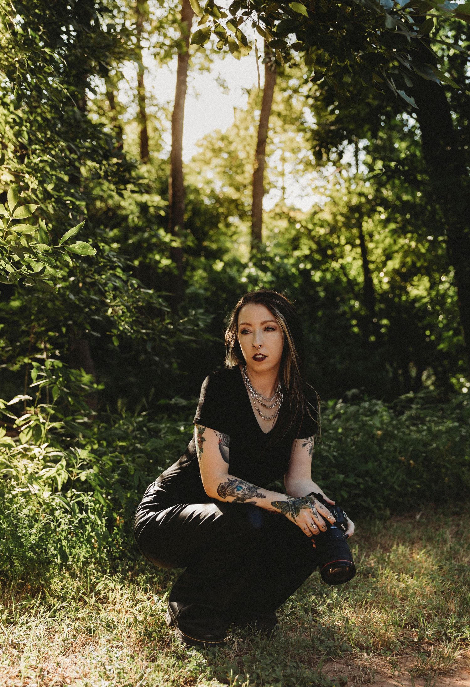 Tattooed Abilene Texas photographer woman in black outfit crouching in lush green forest with sunlight filtering through trees.