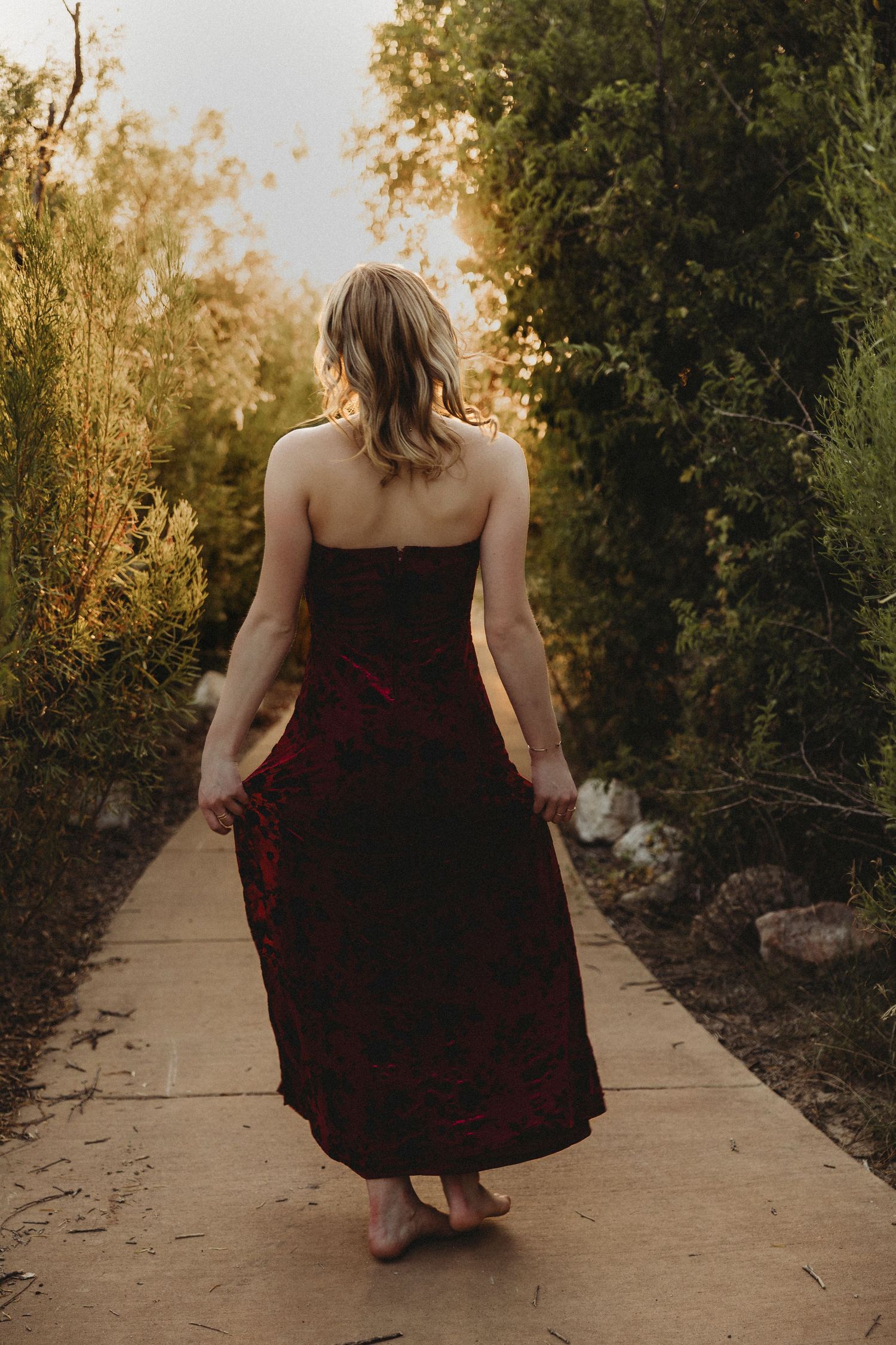 High school senior walking along a tree-lined path in a burgundy dress during a golden hour senior portrait session in Abilene, Texas.