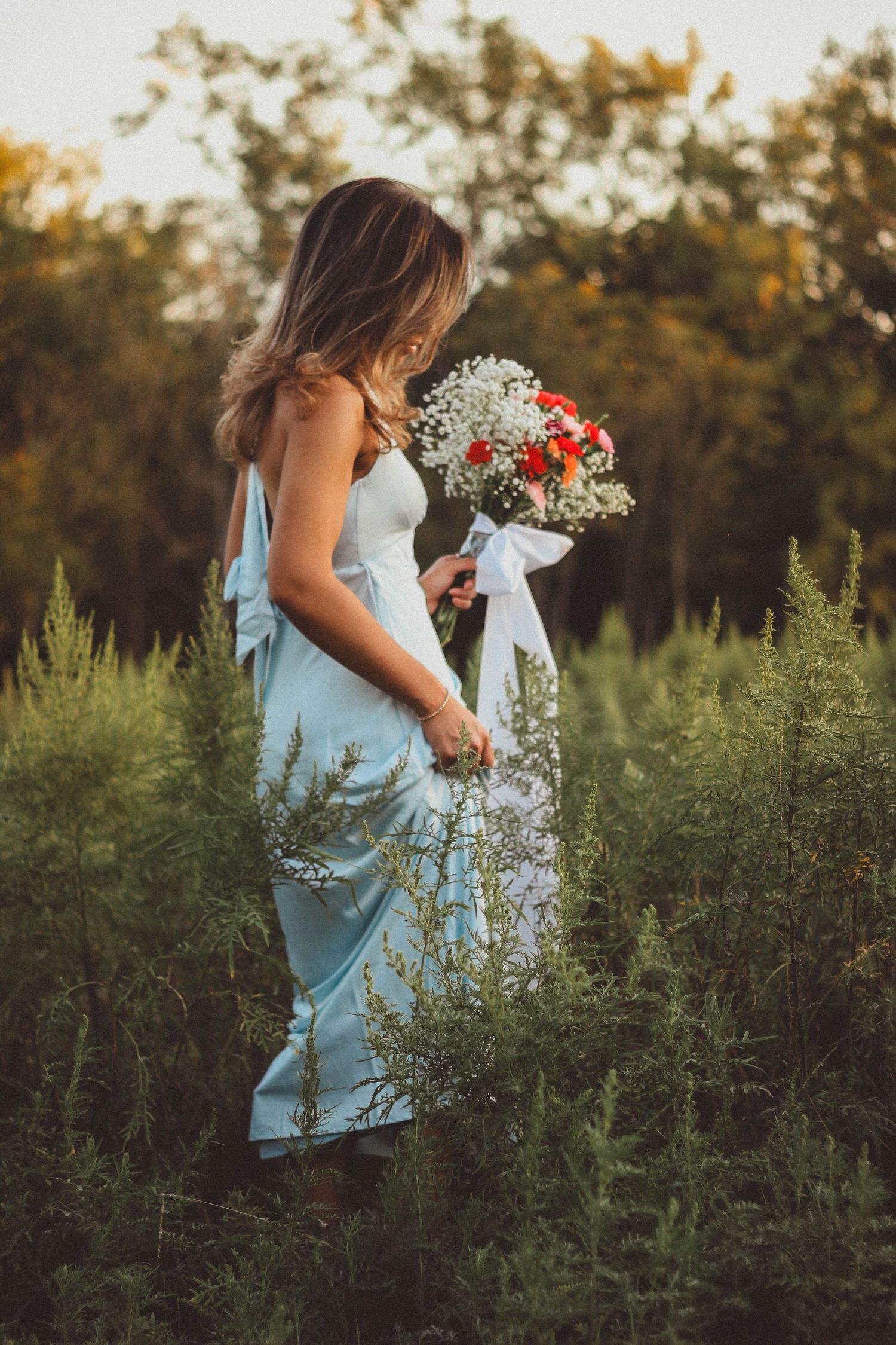 Senior girl in a light blue dress holding a bouquet of flowers in a wildflower field at sunset.
