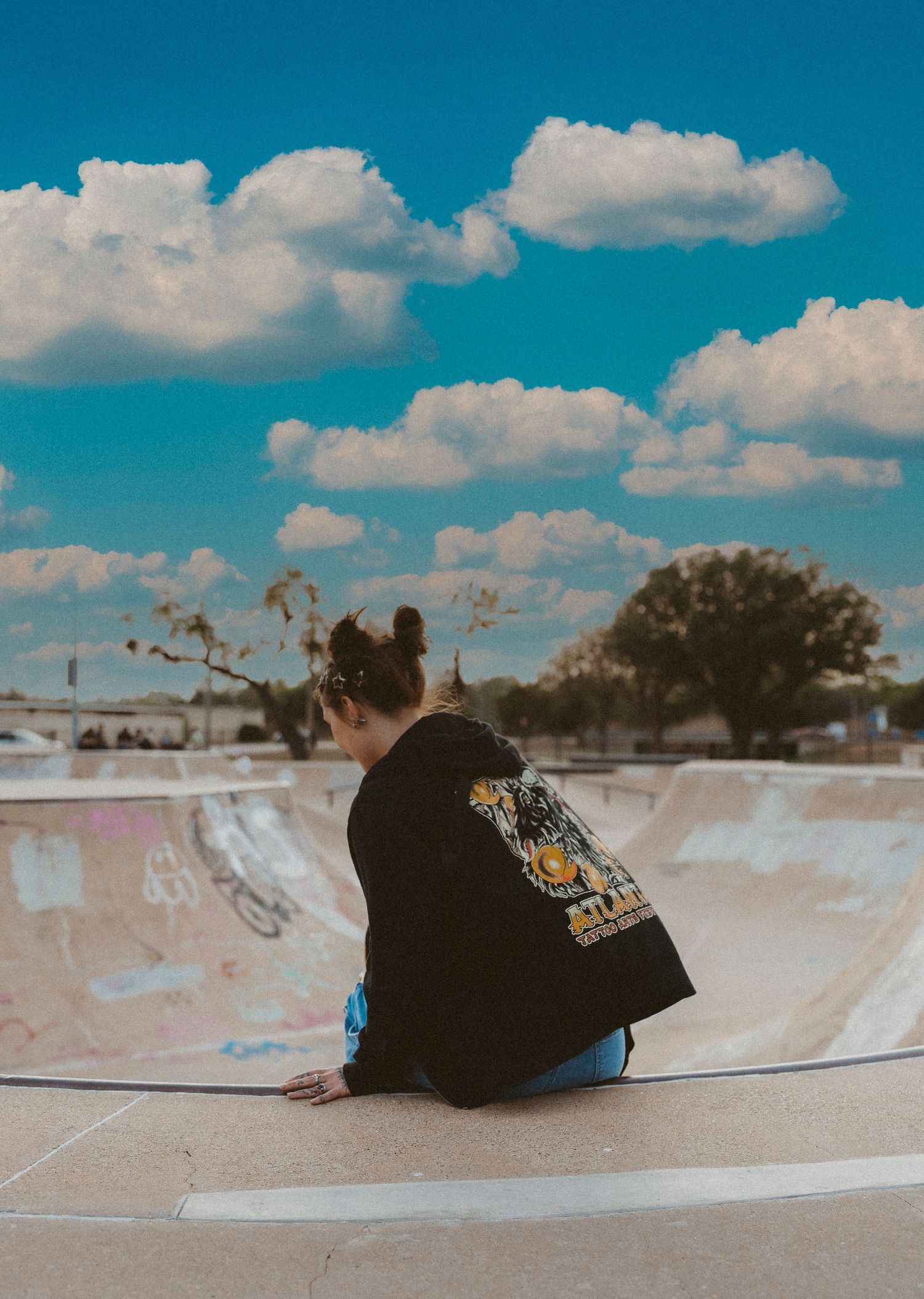 A girl in a black graphic tee sits on a skate ramp at a skatepark in Abilene TX under a vibrant blue sky with fluffy white clouds.
