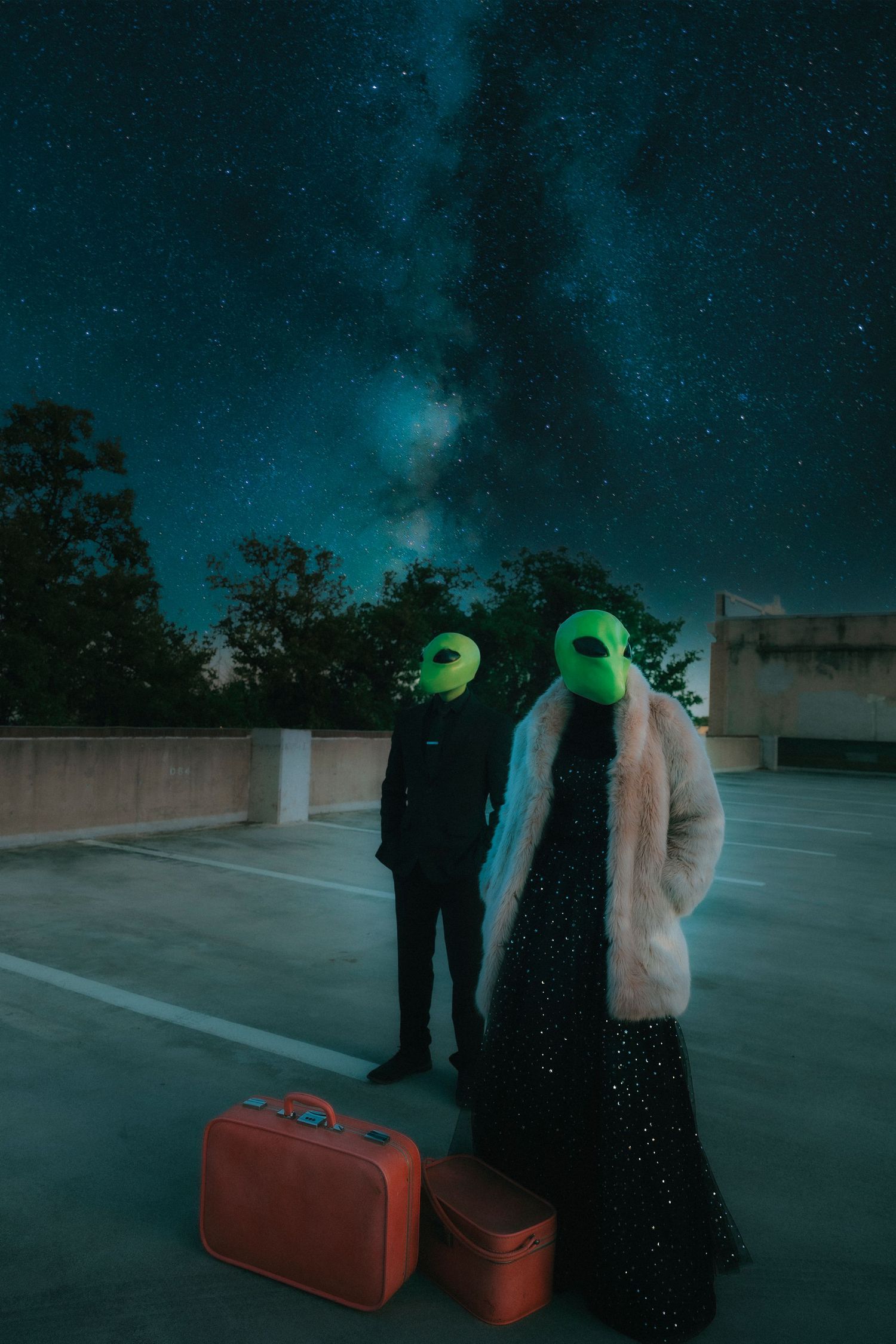 Two people wearing alien masks stand on a rooftop parking lot with red suitcases under a starry night sky.