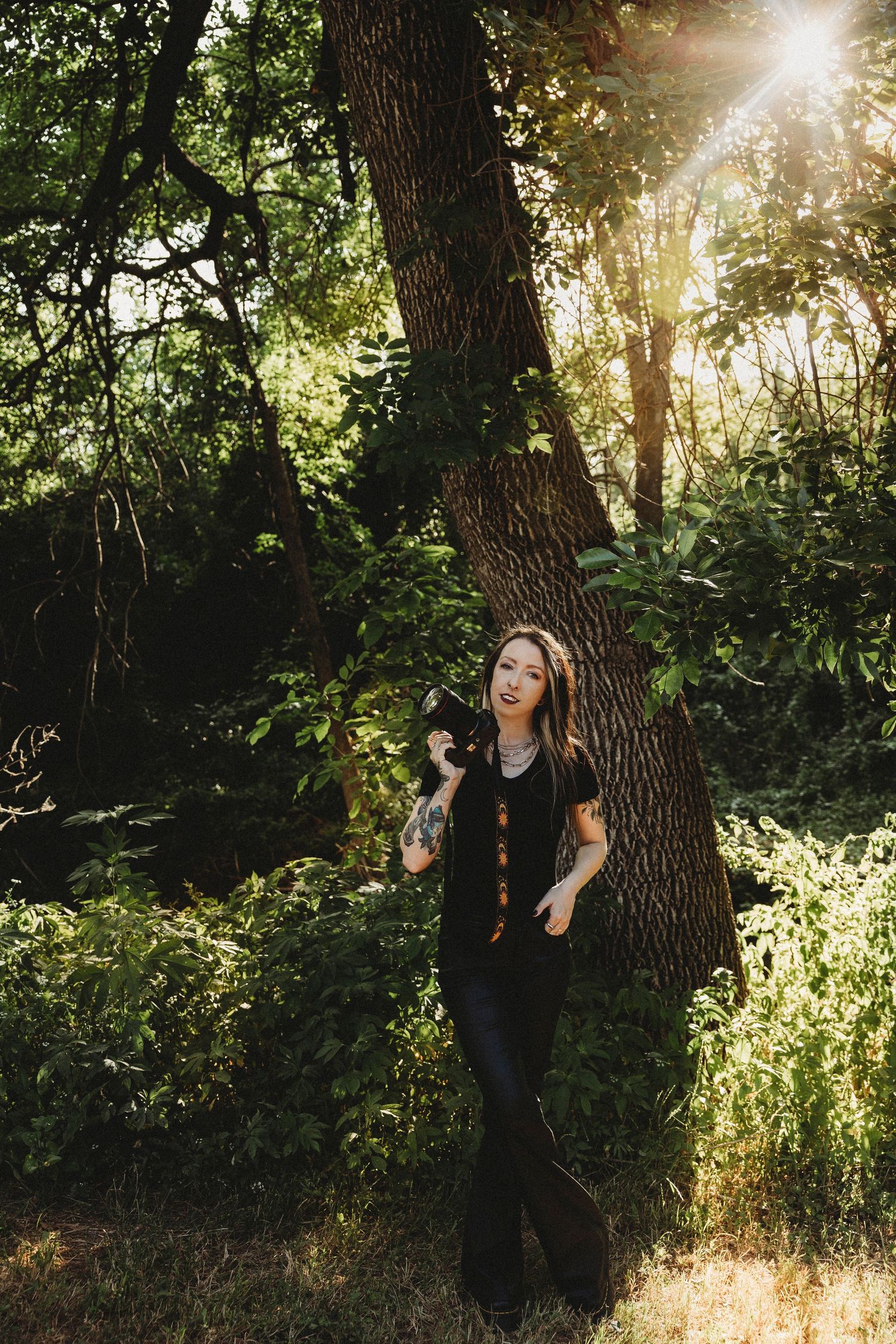 A smiling woman with tattoos holds a camera while leaning against a large tree in a sunlit forest. In Abilene Texas