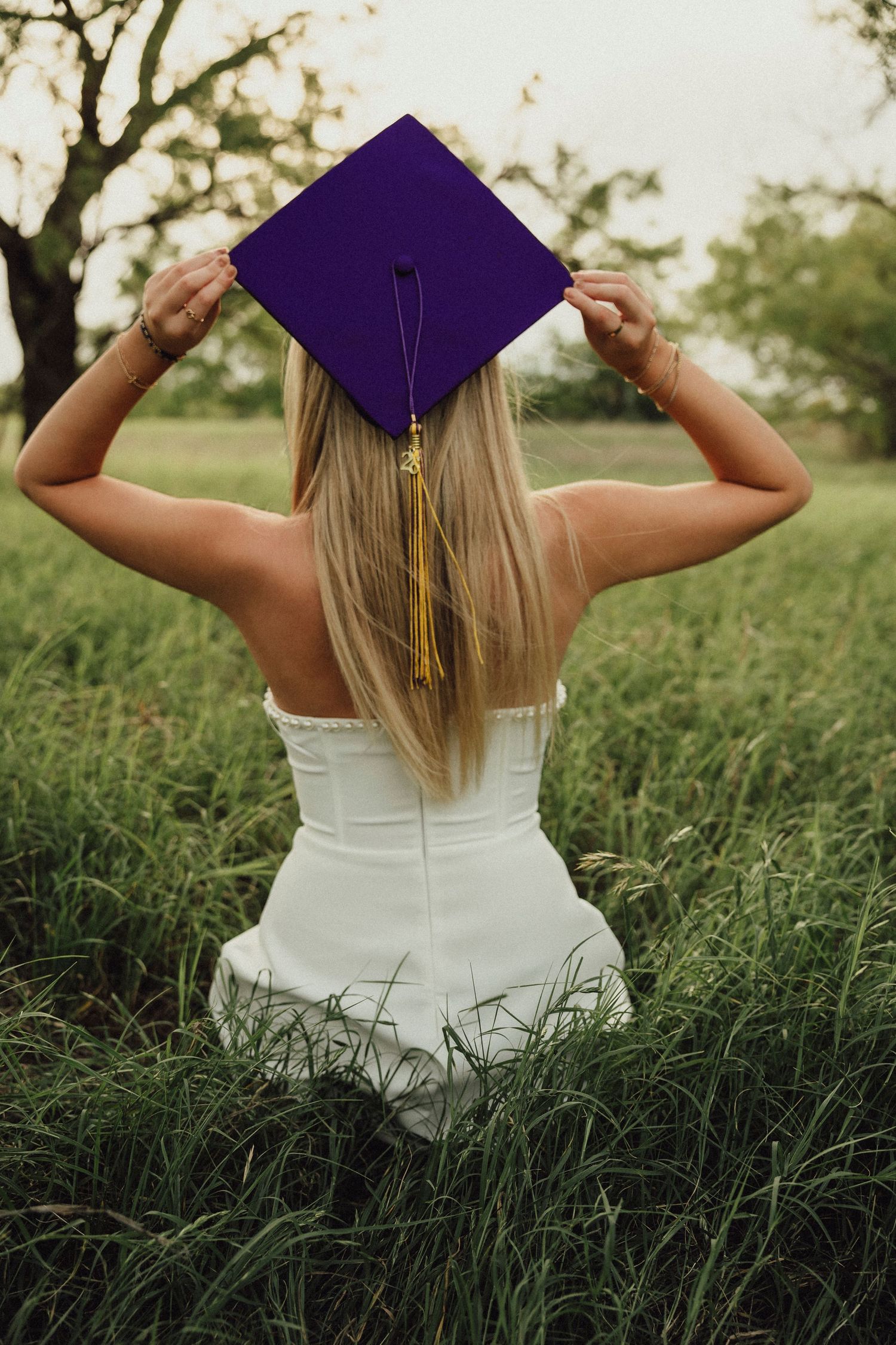 A high school senior in a white dress holds a purple graduation cap with gold tassel while sitting in a lush green field.