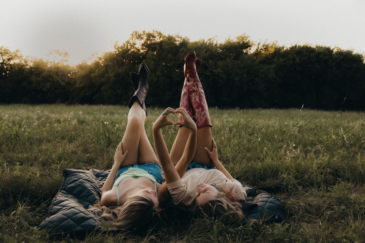 Two teen best friends relax on a blanket with cowboy boots raised in the air during a summer golden hour photoshoot in Abilene, Texas.