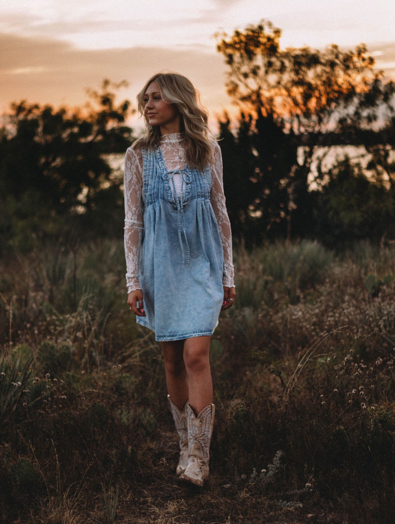 High school senior in a blue dress and cowboy boots walking through a field at golden hour in Abilene, Texas.