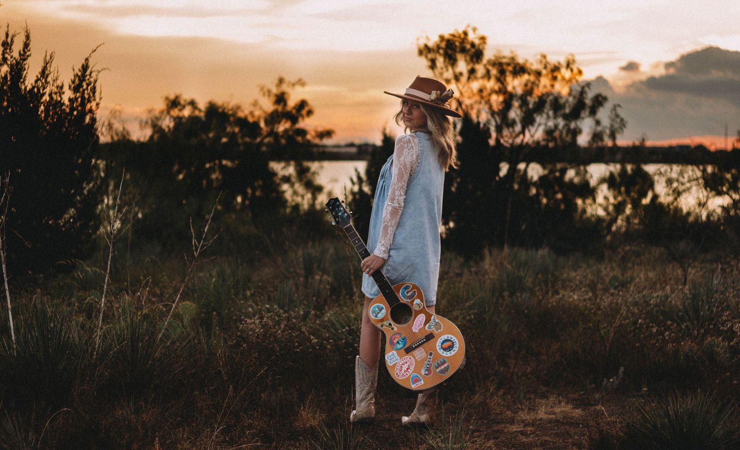 A silhouetted figure stands in a field at sunset holding a guitar while warm golden light illuminates the landscape.