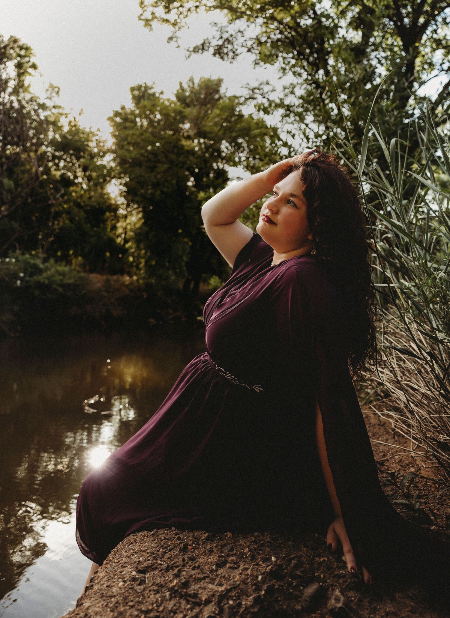 High school senior in a burgundy dress posing beside a peaceful creek during a golden hour senior portrait session in Abilene, Texas.