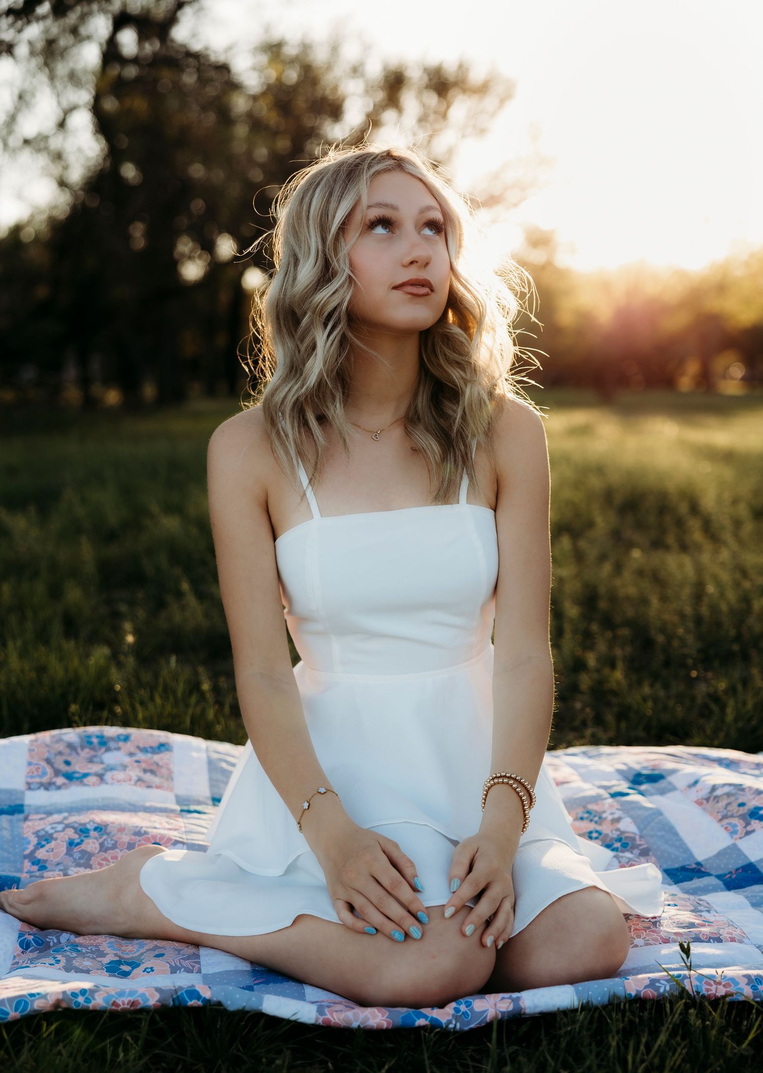 Wylie Senior High School girl in white sundress sitting cross-legged on floral quilt in sunlit green field at golden hour.