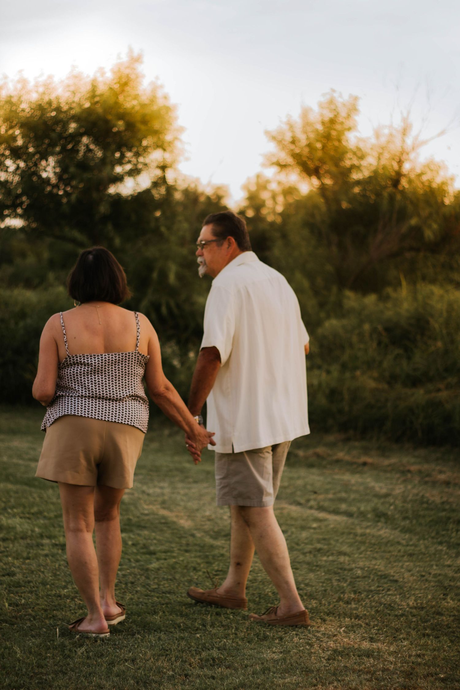 Older couple holding hands and walking barefoot on green grass at sunset with lush trees in the background.