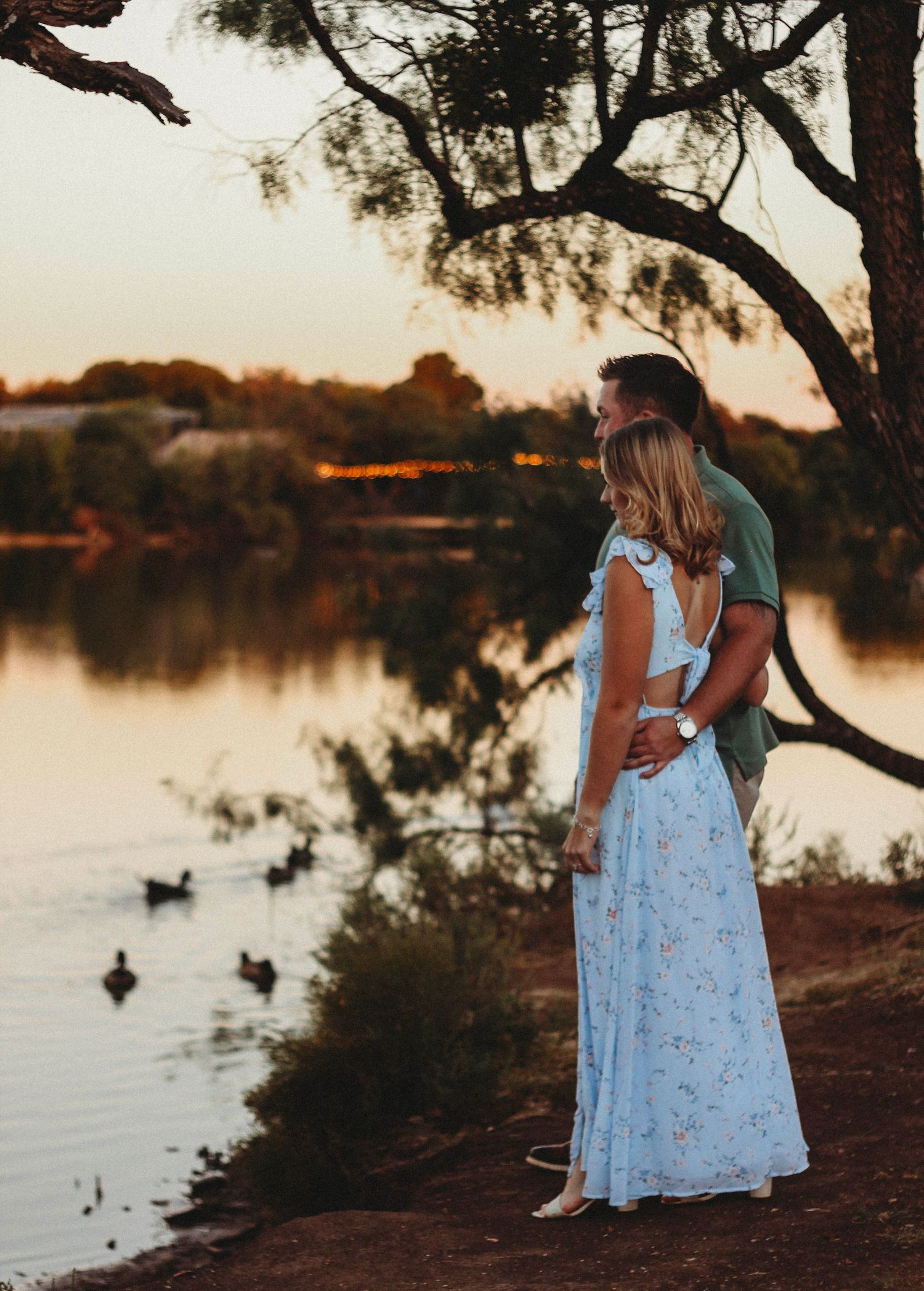 Couple embracing by a tranquil lake at sunset, surrounded by trees and ducks, woman in floral blue maxi dress.