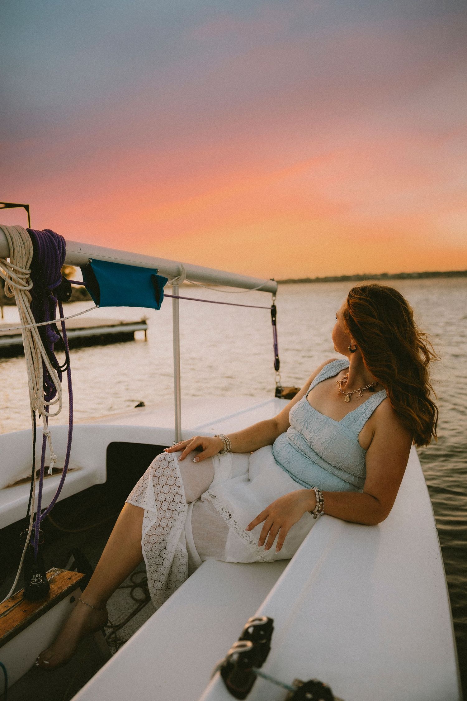 Abilene TX Senior relaxing on a sailboat at sunset, wearing a light blue top and white skirt, gazing at colorful sky.