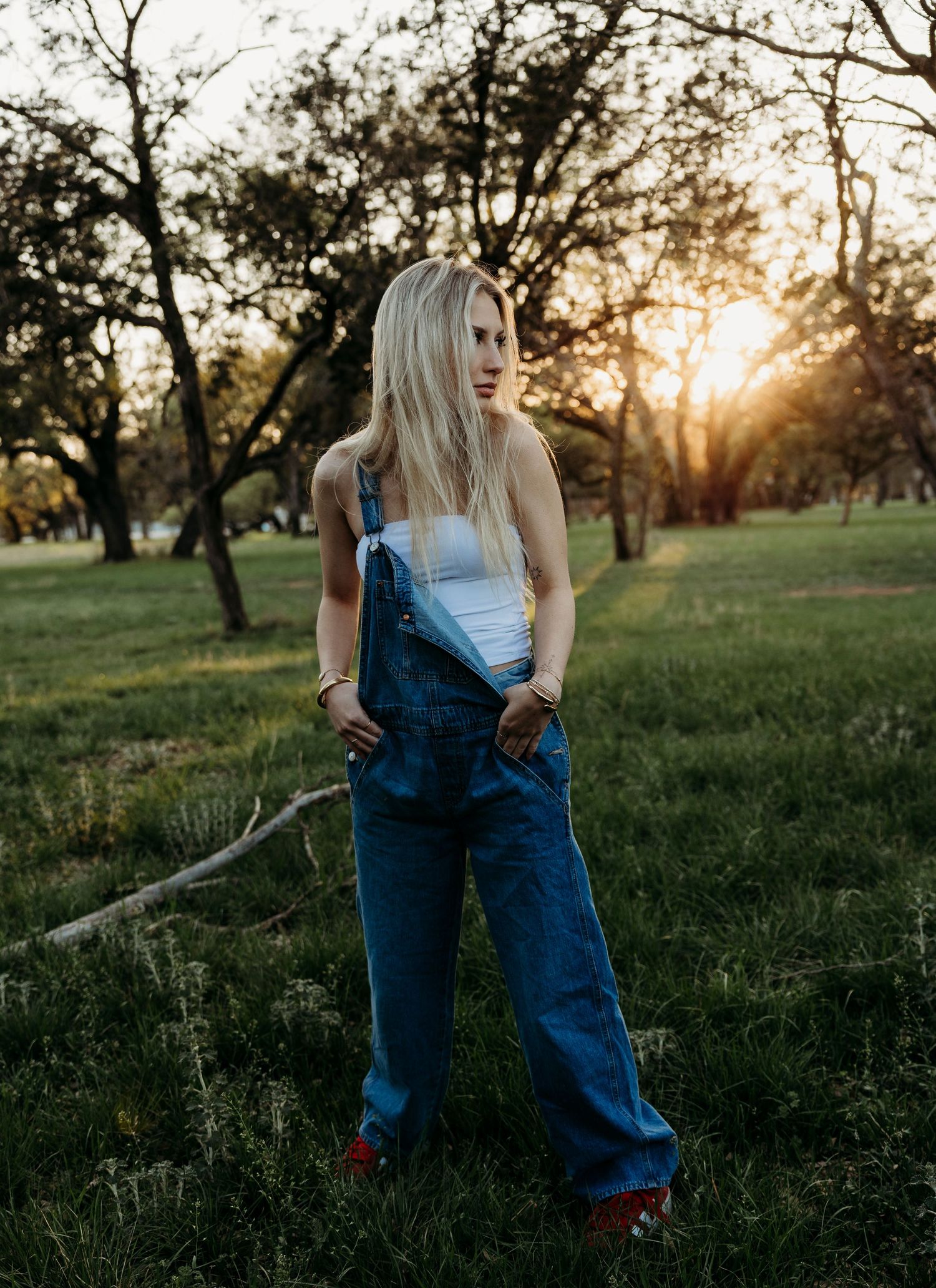 Senior girl  in denim overalls and white top stands in a grassy field at golden hour sunset.