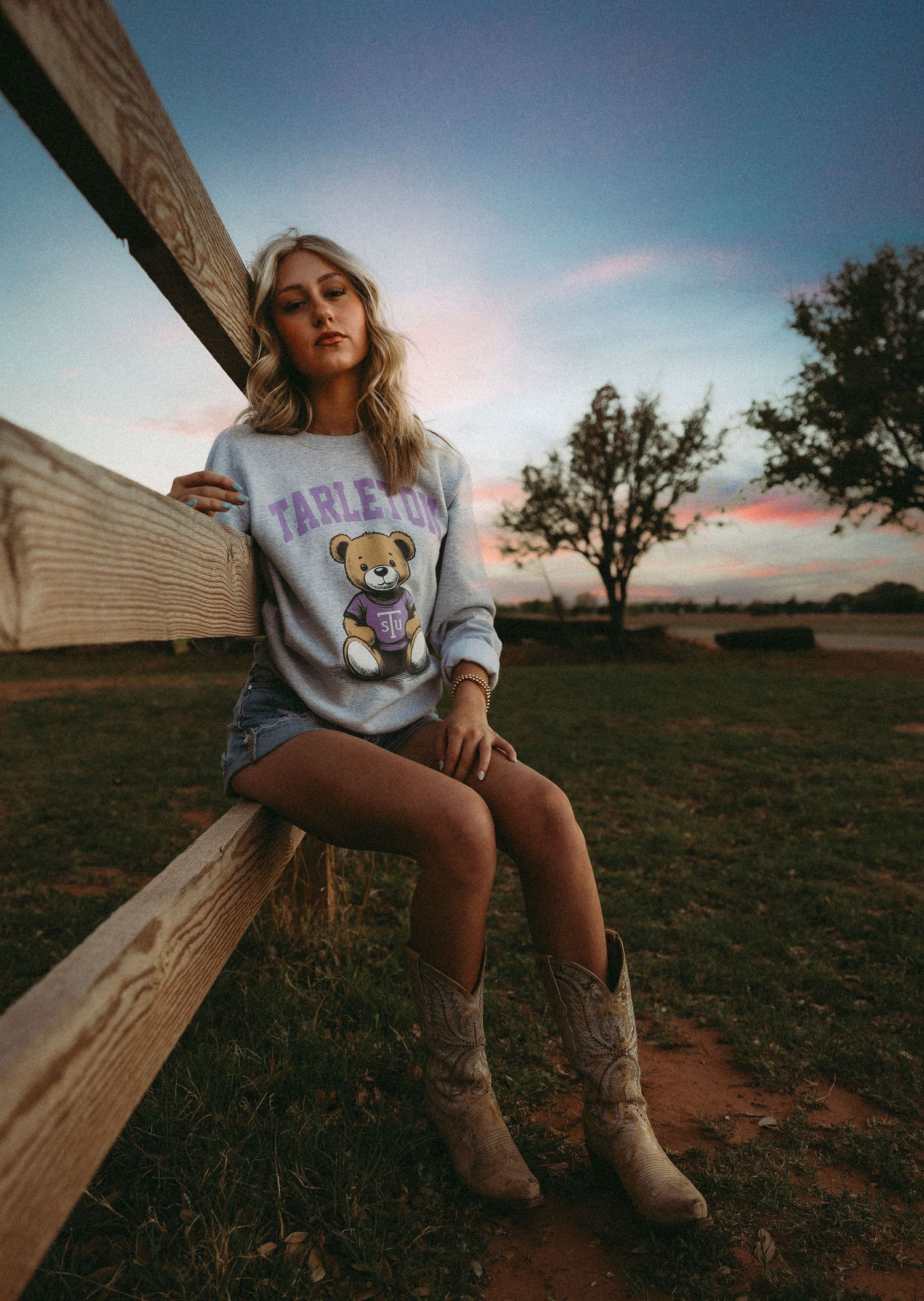Senior girl  in Tarleton sweatshirt and cowboy boots sitting on a fence at sunset on a farm.