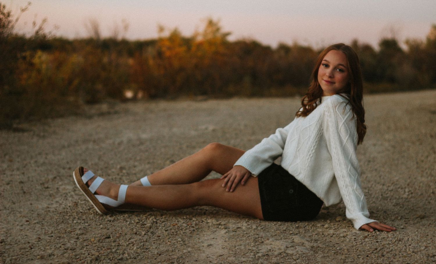 Senior girl in a white sweater and black shorts sitting on a gravel path at golden hour with autumn foliage behind her.