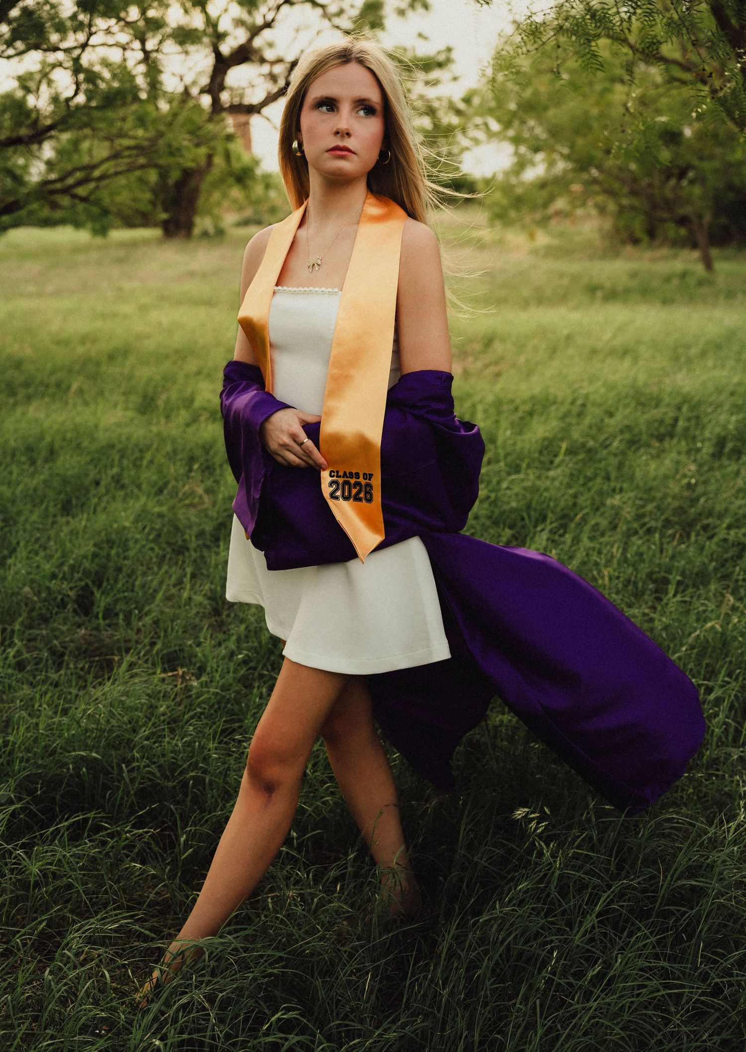 High School Graduate in white dress with purple robe and gold Class of 2026 sash posing outdoors in green field.